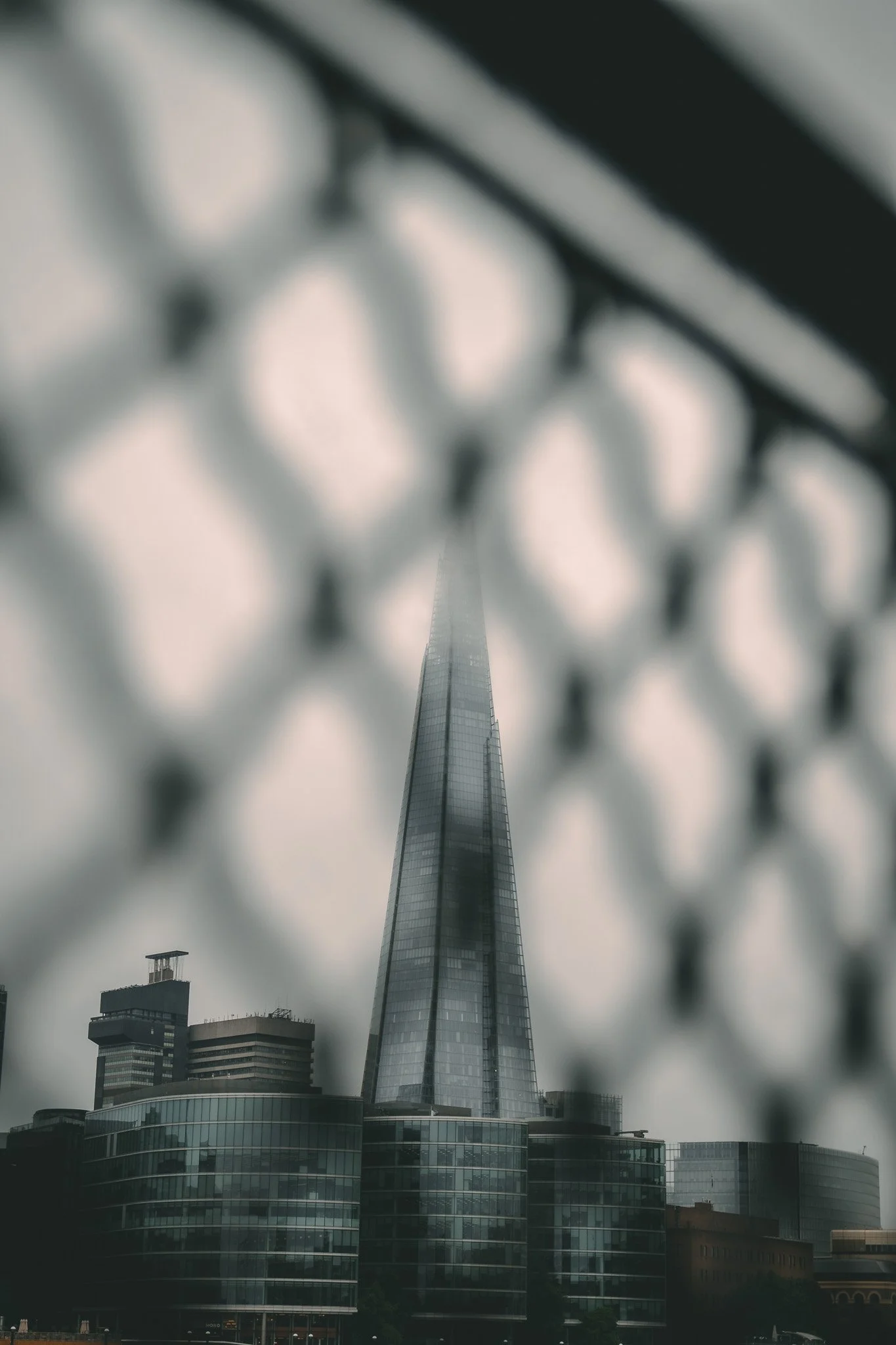 Tall skyscraper viewed through a chain-link fence with a cloudy sky in the background.