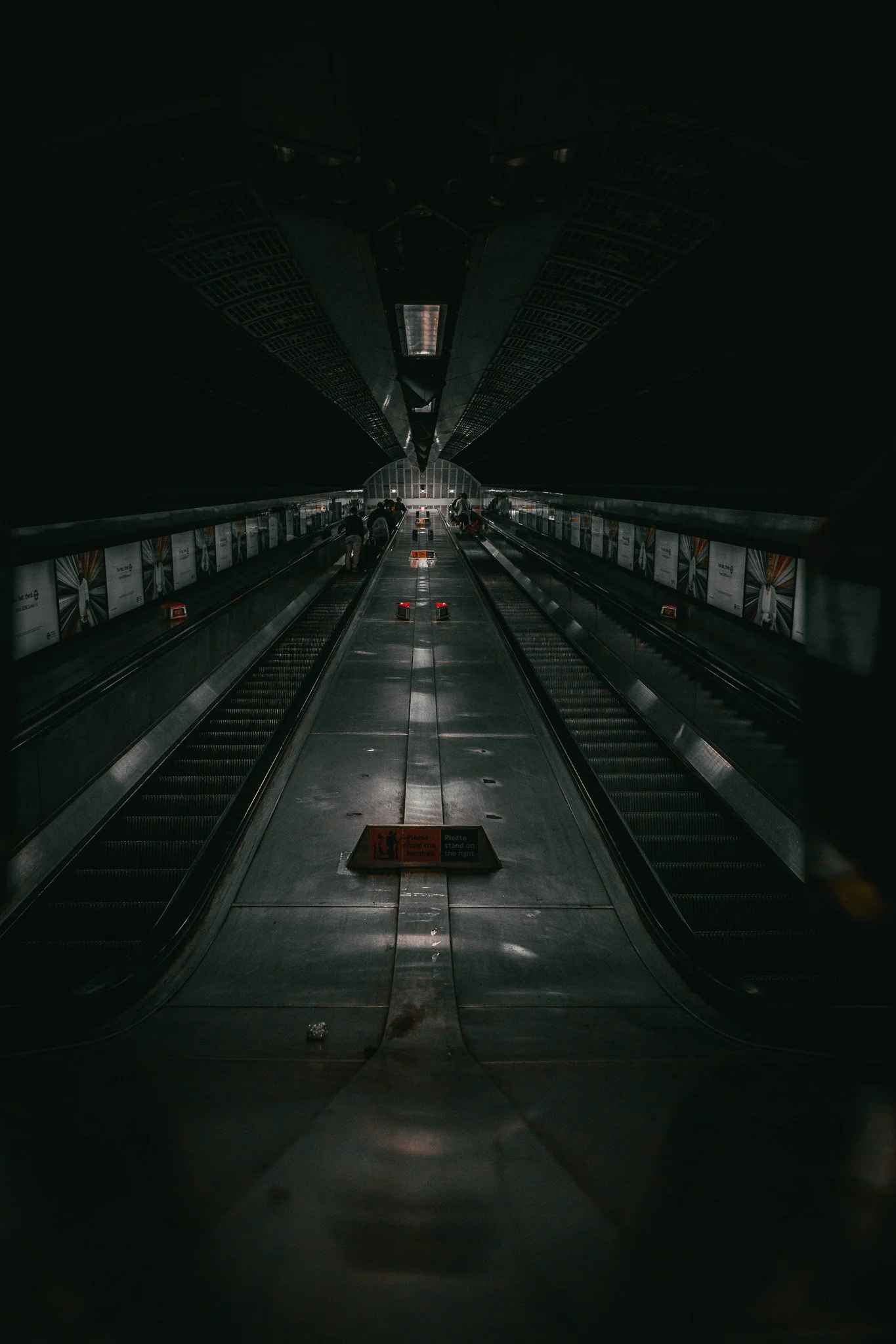 An empty and dark moving walkway inside a modern subway station with minimal lighting and a few people in the distance.