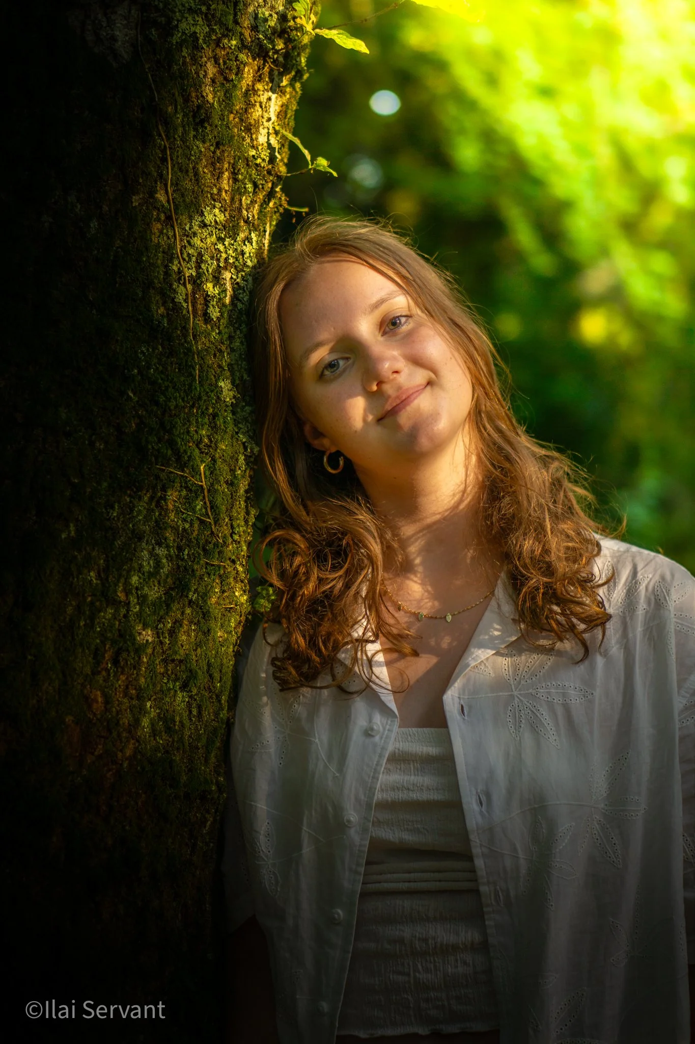 A young woman with curly reddish hair and earrings smiling in a forest beside a moss-covered tree, with sunlight filtering through the green foliage.