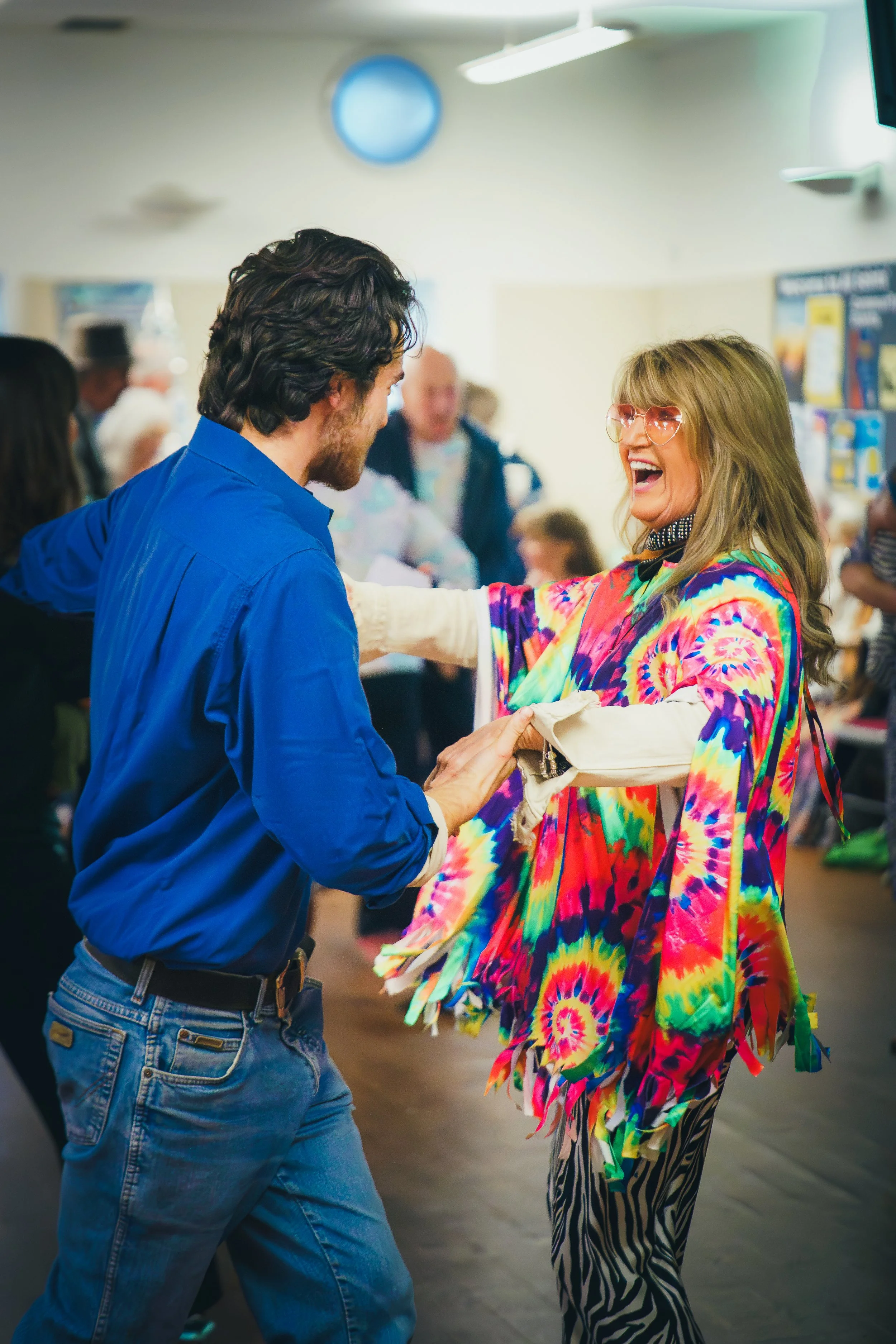 Two people dancing and smiling in a crowded indoor space. The woman on the right is wearing glasses, a colorful tie-dye jacket, and zebra-striped pants. The man on the left is wearing a blue shirt and jeans.