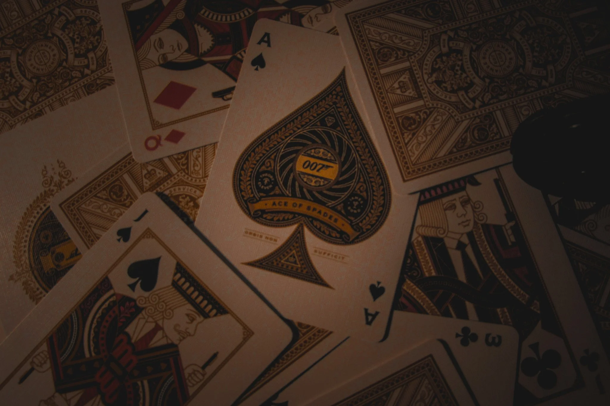 A close-up of playing cards, including the Ace of Spades, Queen of Diamonds, Ace of Hearts, and 3 of Clubs, spread out over a surface with other cards and casino-themed designs visible.