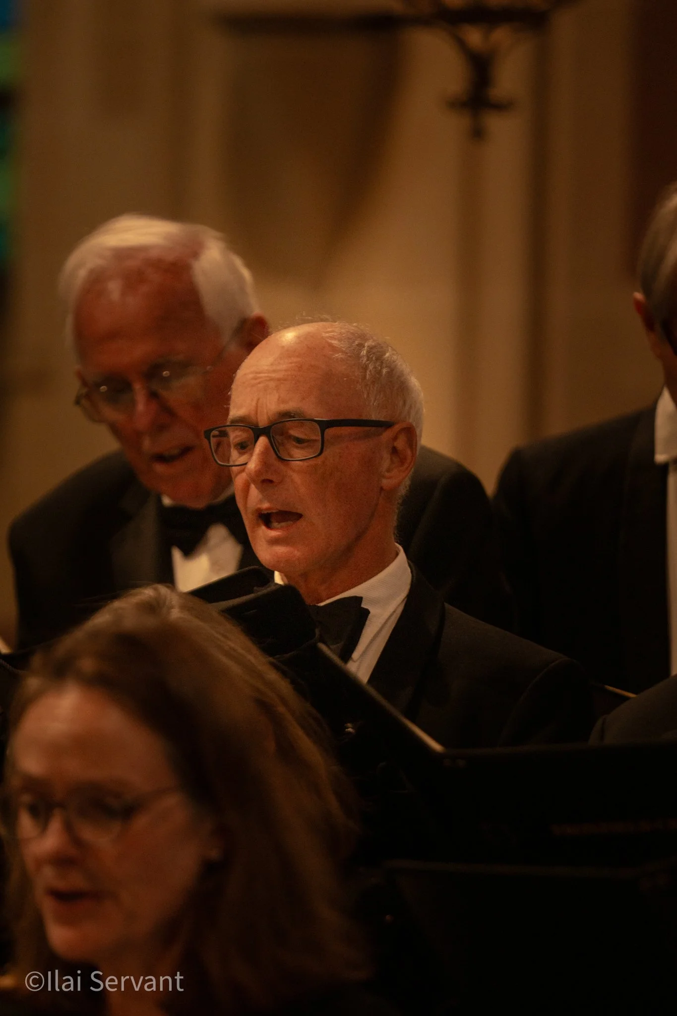 A group of seniors in tuxedos singing together at a piano in a dimly lit setting.