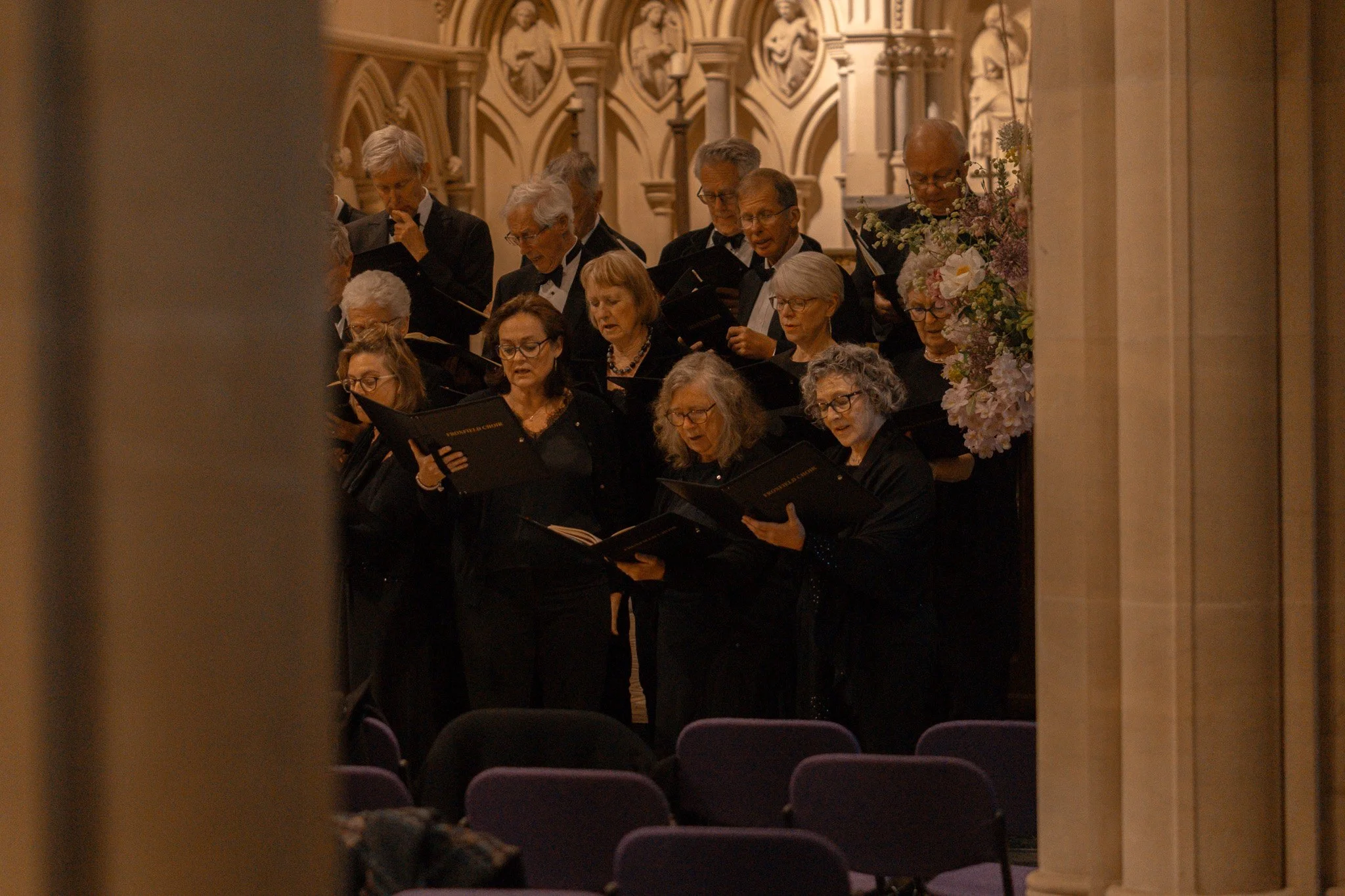 A choir of older adults singing and holding sheet music, standing in a church with ornate architecture and floral arrangements.