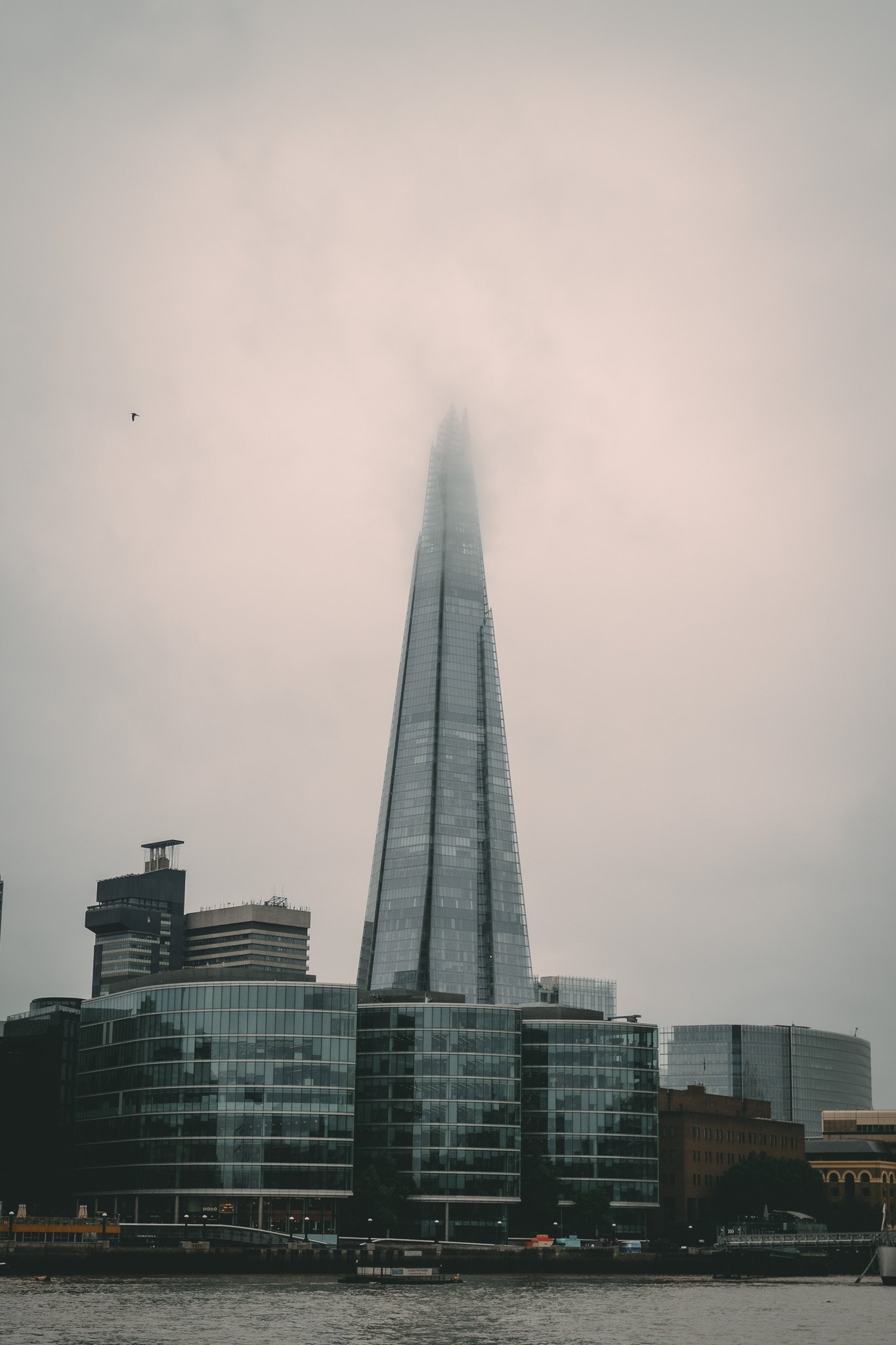 A tall skyscraper shrouded in fog, with a few shorter buildings at its base and a body of water in the foreground.