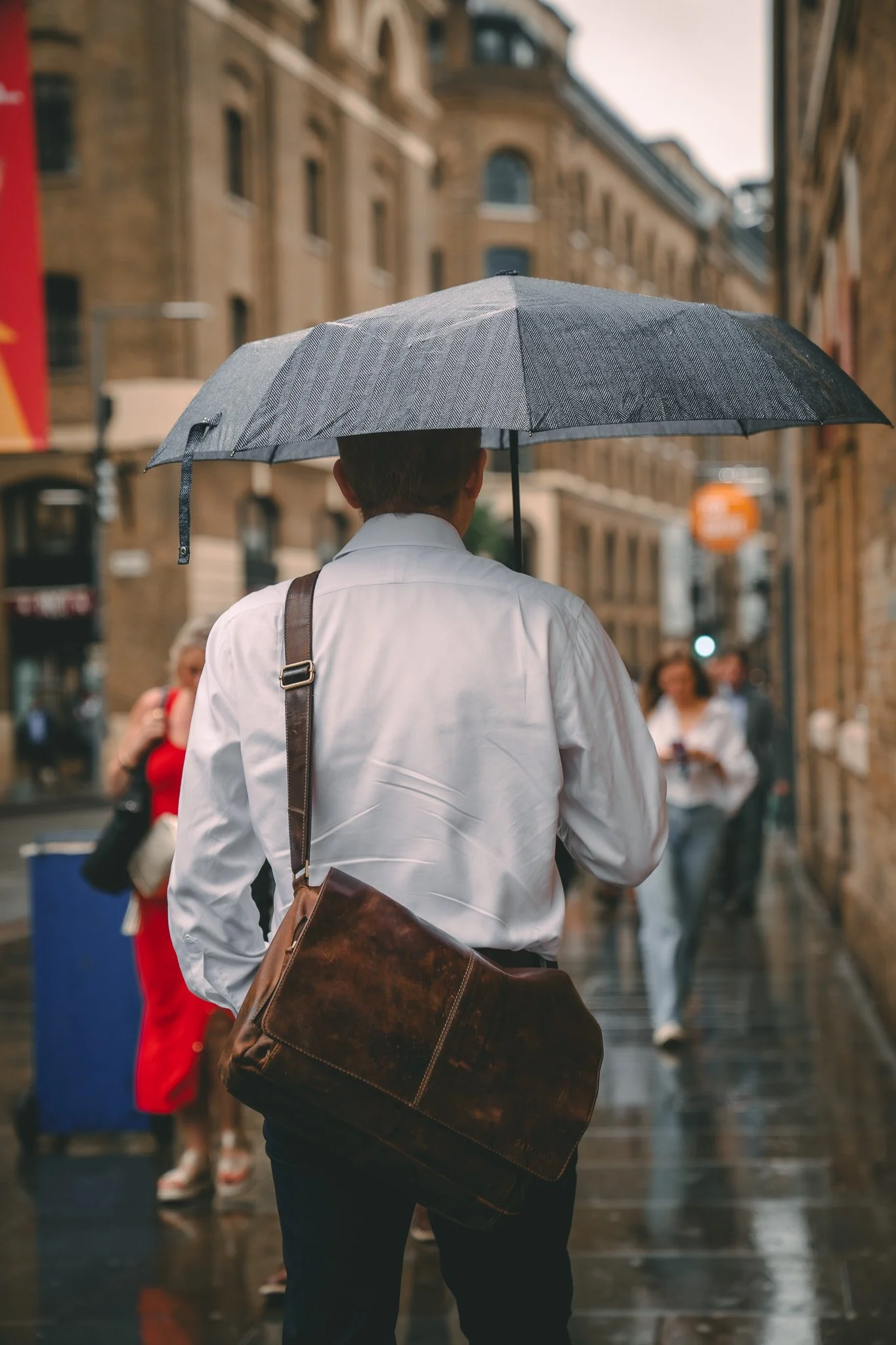 A man with a brown leather shoulder bag holding a grey umbrella walking on a wet city street on a rainy day, with blurred pedestrians in the background.