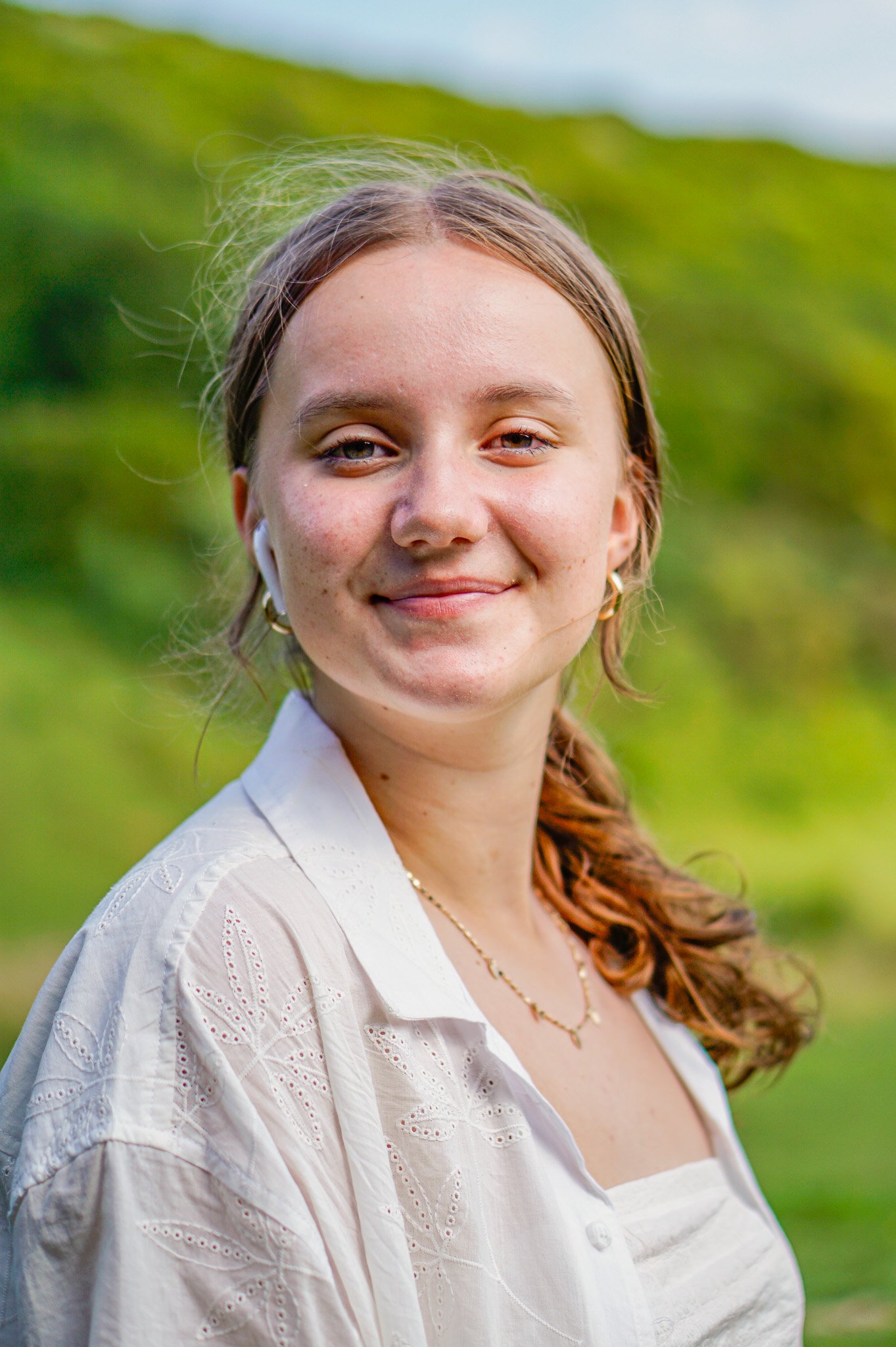A young woman with light skin and long, reddish-brown hair styled in a loose ponytail, smiling at the camera outdoors in a green field.