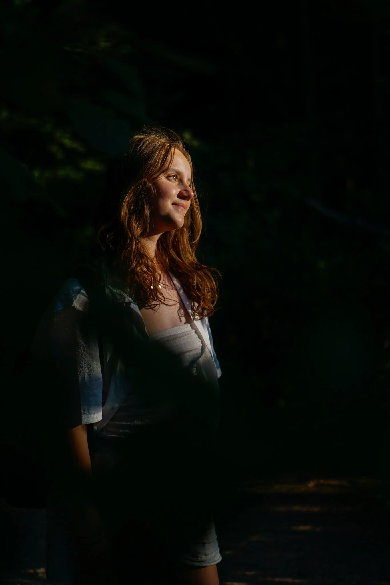 A young woman with long red hair standing outdoors in the sunlight, surrounded by dark trees, gazing into the distance with a peaceful expression.