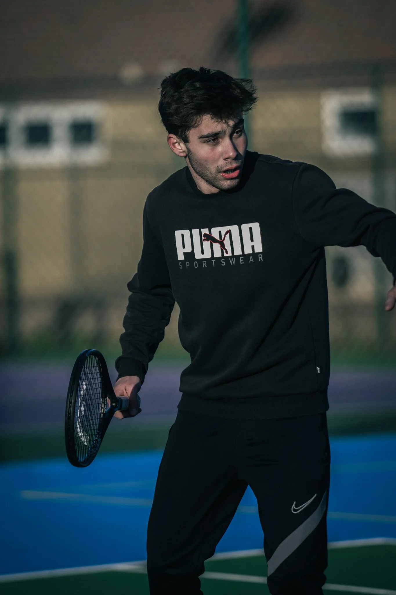 Young man in black Puma sportswear holding a tennis racket on an outdoor tennis court