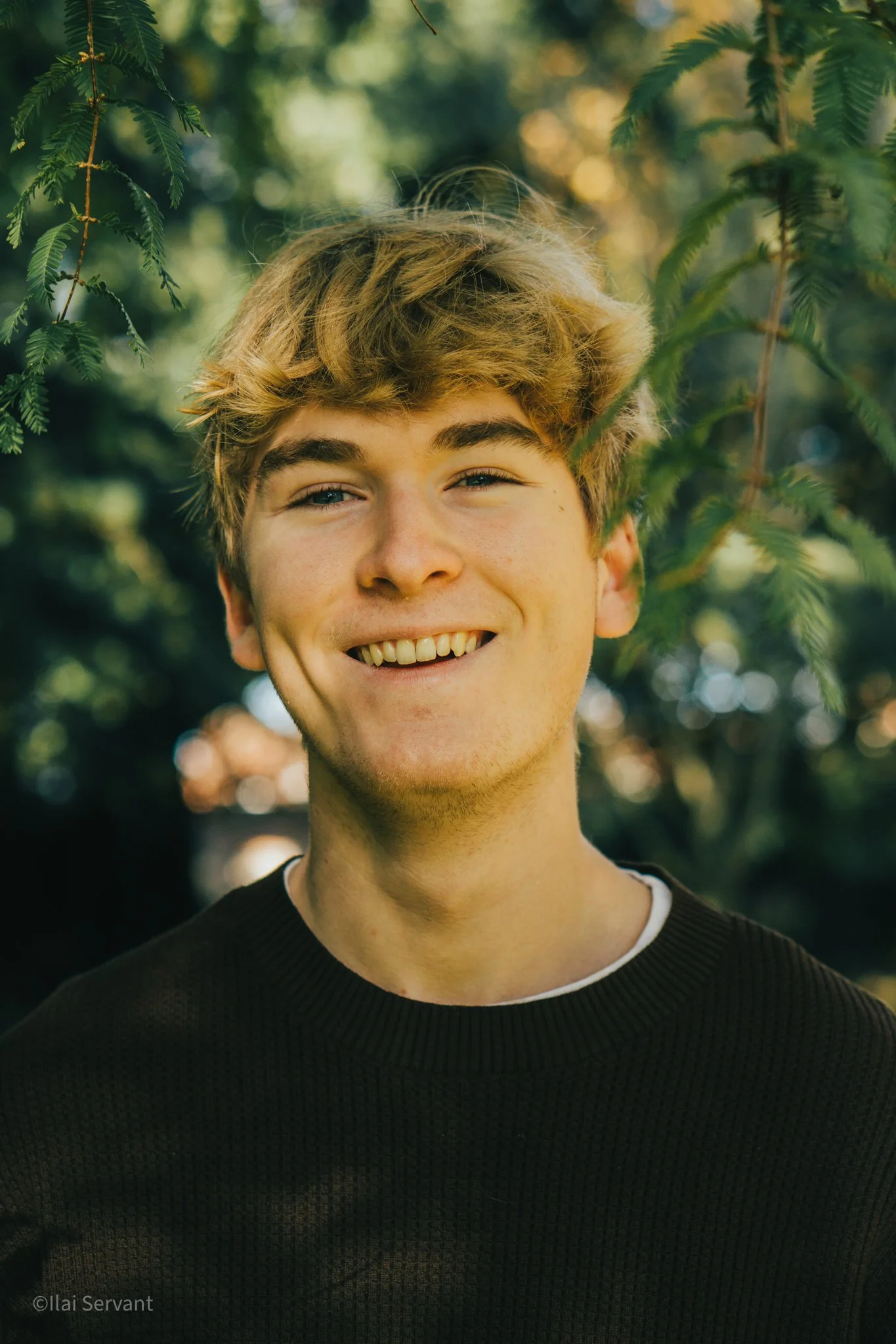 A young man with curly blonde hair smiling outdoors with green trees in the background.