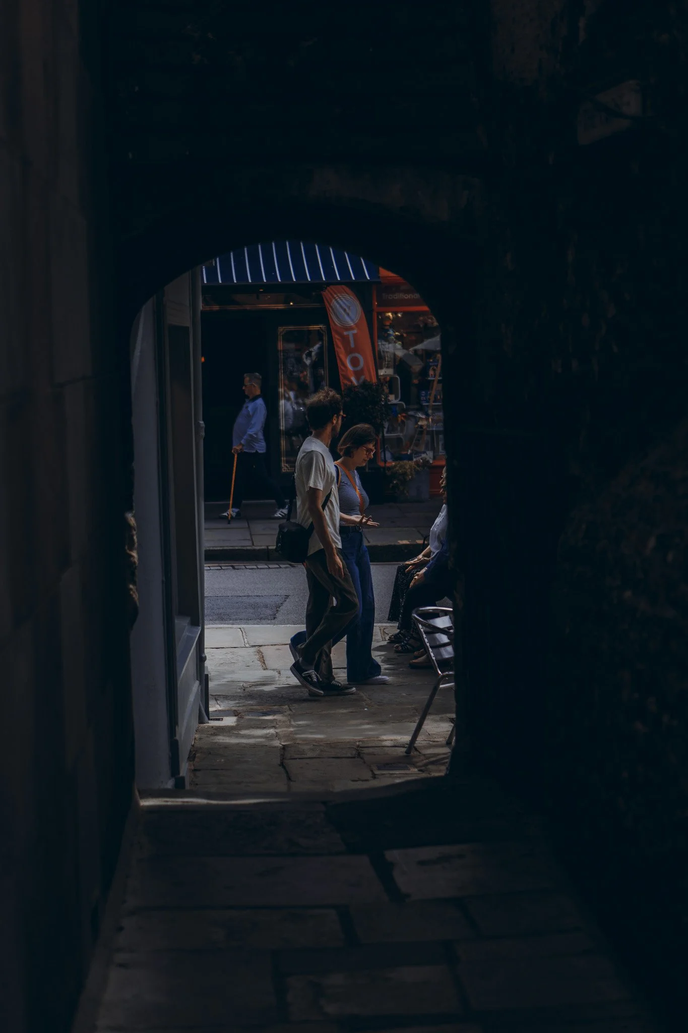 View through a dark archway of people on a city sidewalk, some sitting on chairs, others standing, with a person using a phone, and pedestrians walking in the background.