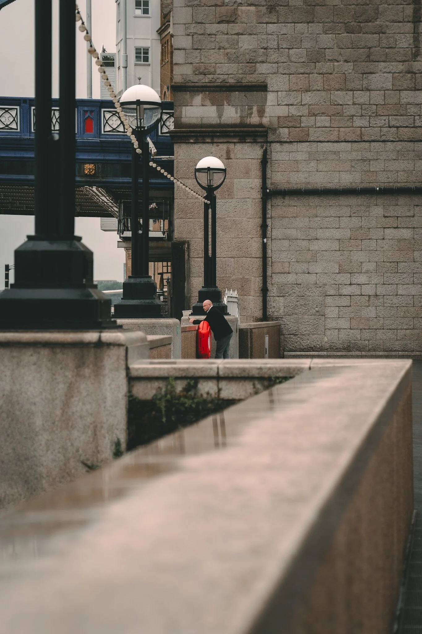 A man in a black jacket and gray pants is leaning over a bright red fire hydrant against a large stone building wall, under a row of black street lamps with round light fixtures, near a blue bridge.