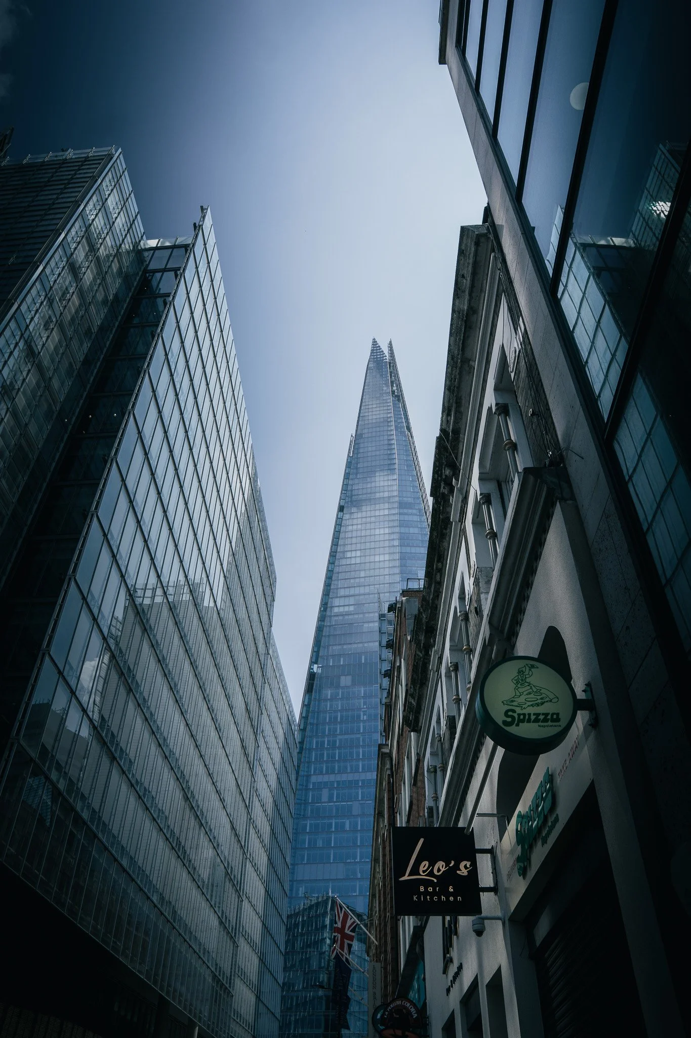 A city street view looking up at skyscrapers, including the Shard in London, with signs for Leo's bar and kitchen, Spizza restaurant, and other buildings.