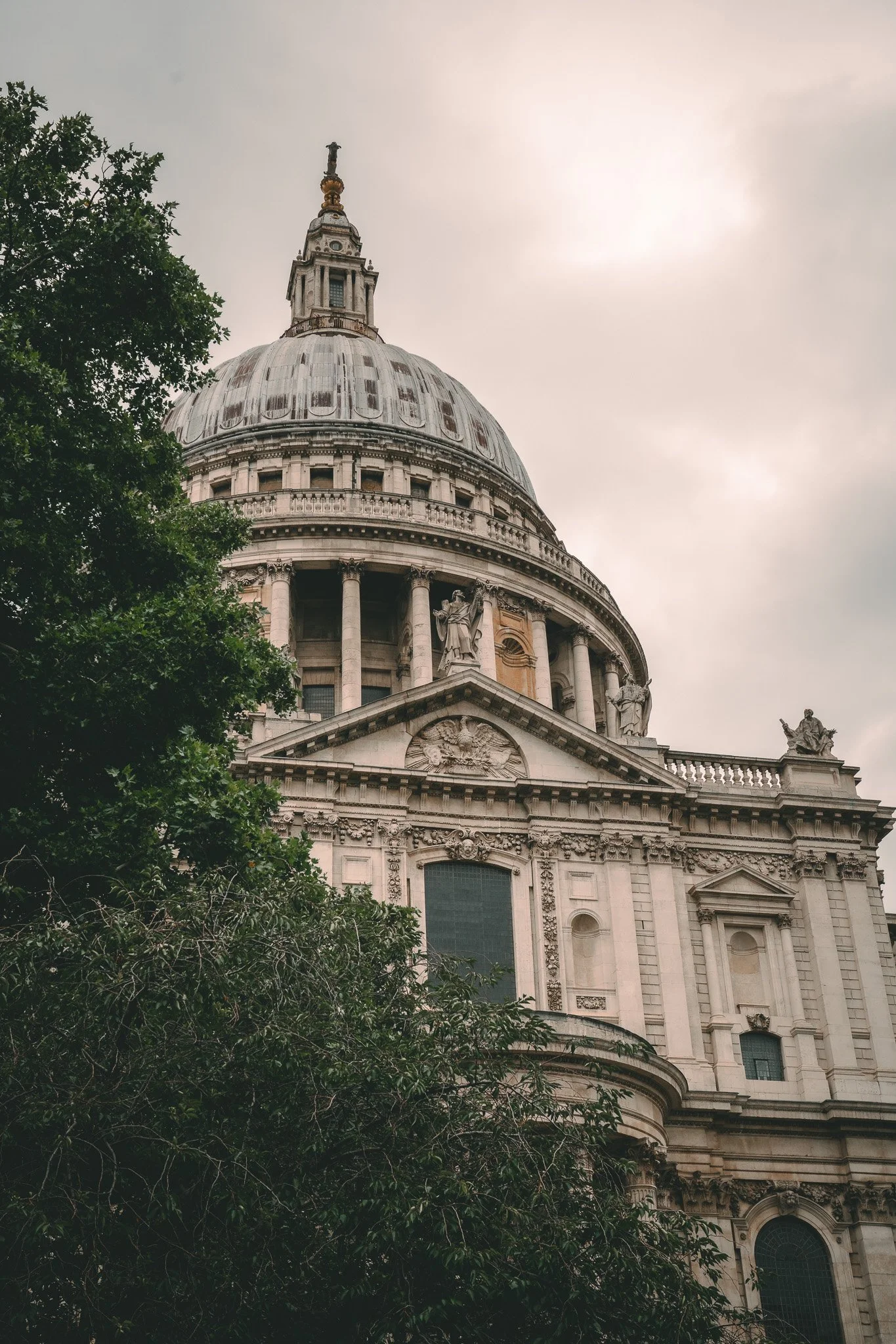 The dome of a historic building, possibly a cathedral or government building, with ornate architectural details, columns, statues, and a weathered roof, partially obscured by green trees, under a cloudy sky.