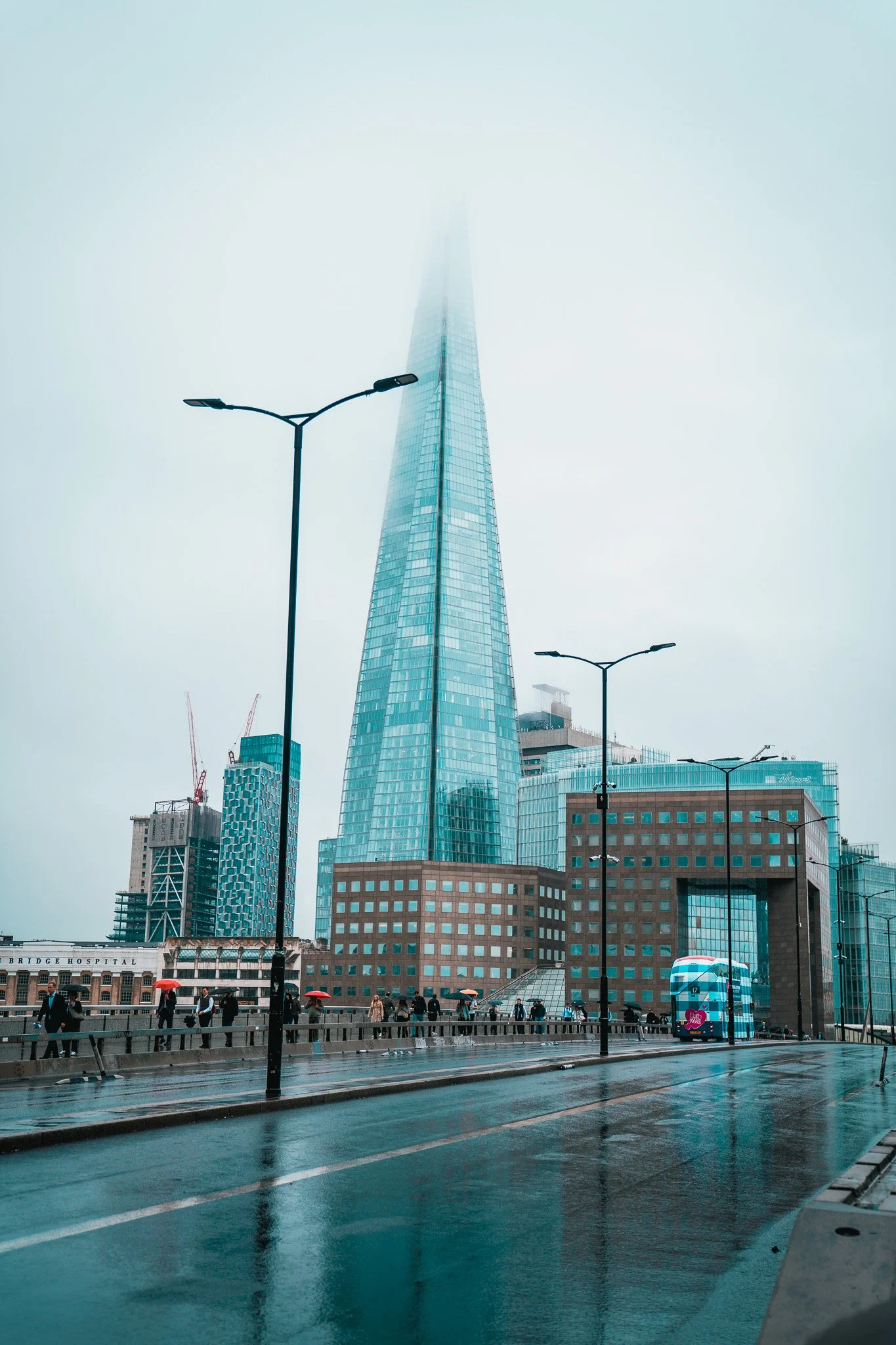 Cityscape featuring the sky with fog covering the top of a tall, modern glass skyscraper, with people holding umbrellas walking on a rainy street.