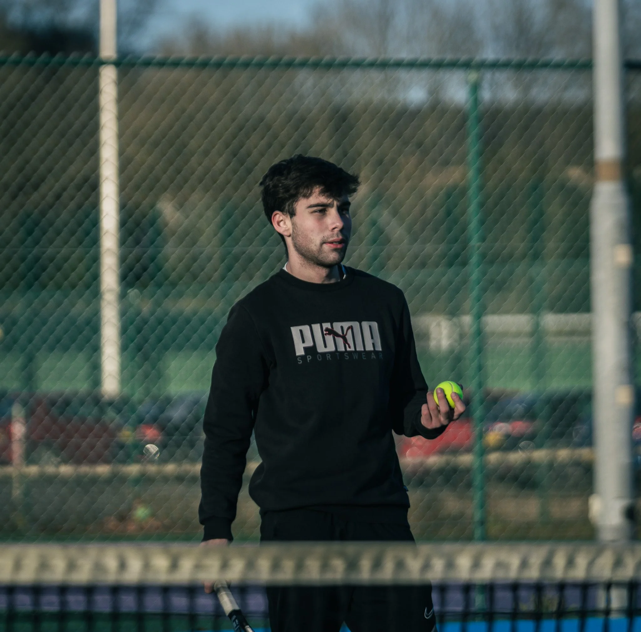 A young man with dark hair and a serious expression holding a bright yellow tennis ball on a tennis court with a chain-link fence in the background.