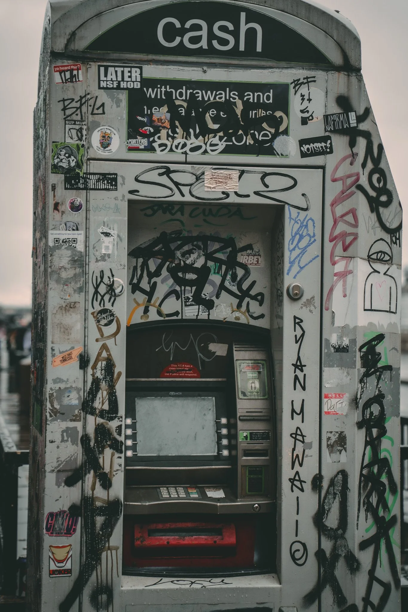 Graffiti-covered ATM machine with a green 'cash' sign on top and various stickers, tags, and scribbles on the surrounding surface.