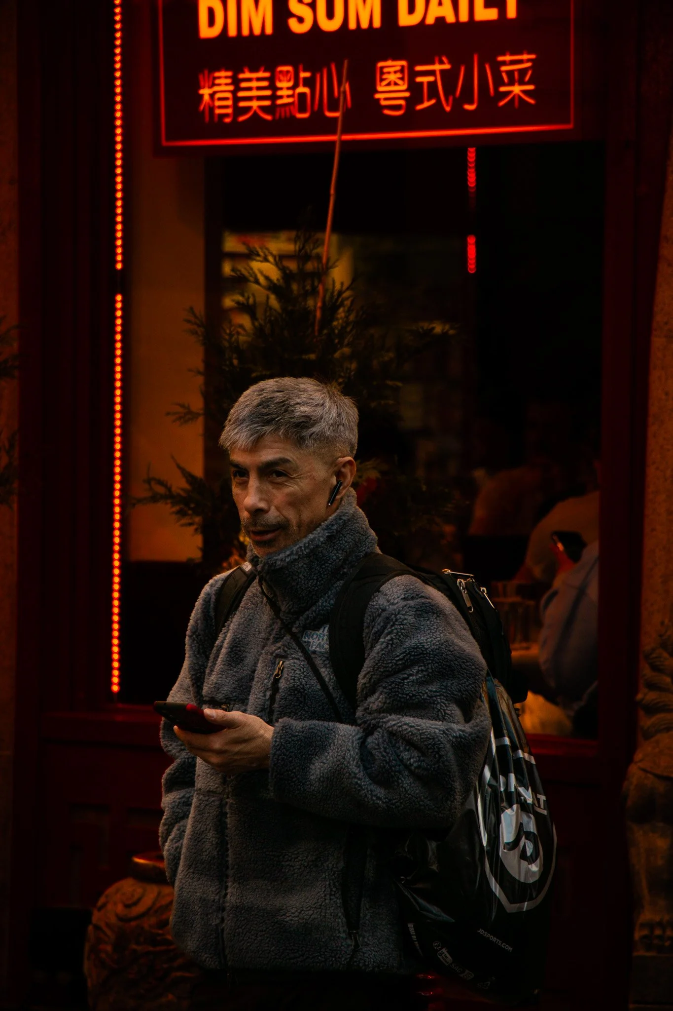 Man in gray fleece jacket with backpack standing outside restaurant with neon sign in Chinese characters and English text that reads "Dim Sum Deli."