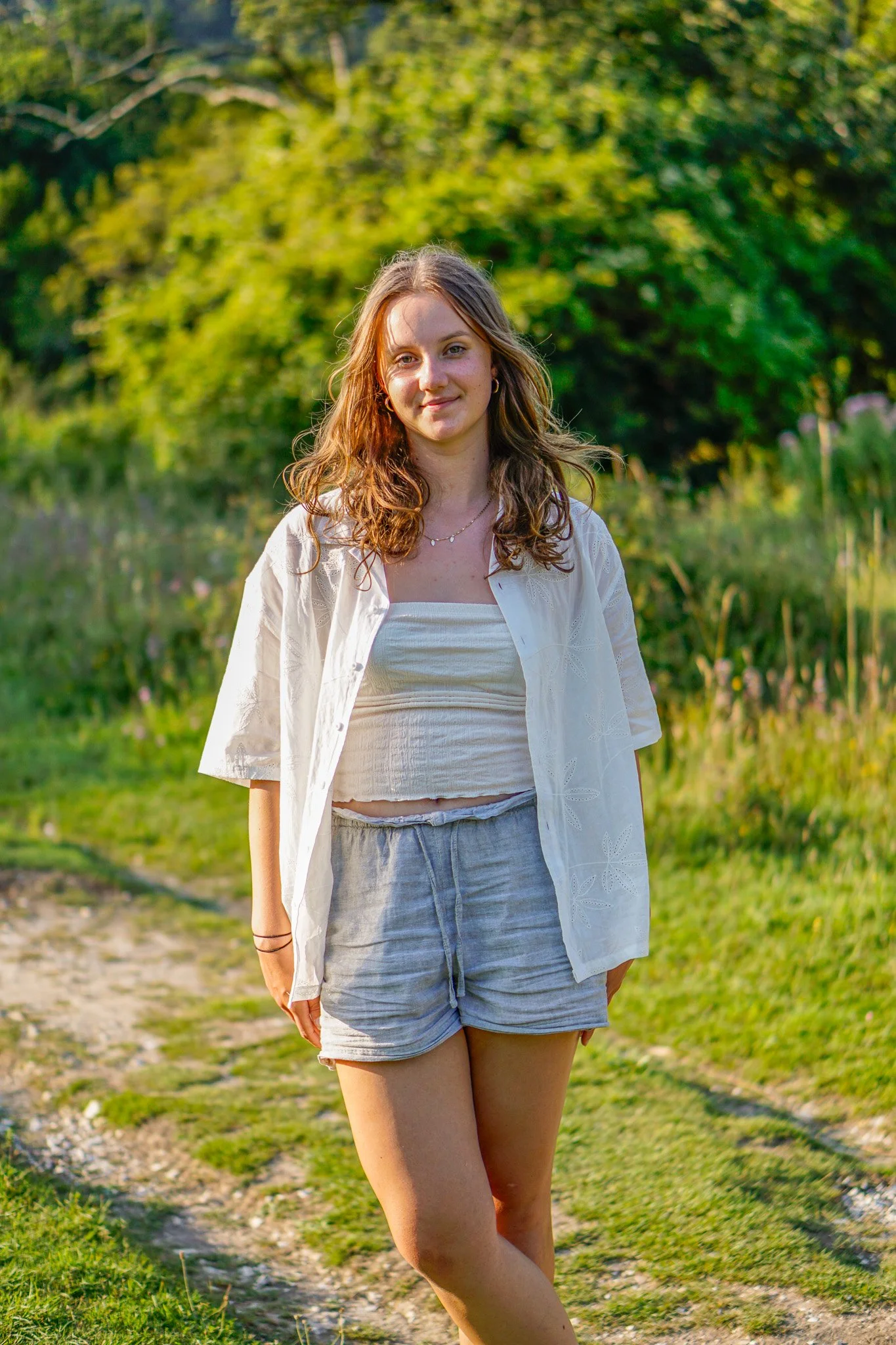 Young woman with curly brown hair wearing a white top, light gray shorts, and a white open shirt, standing outdoors on a grassy path with trees and bushes in the background during the daytime.
