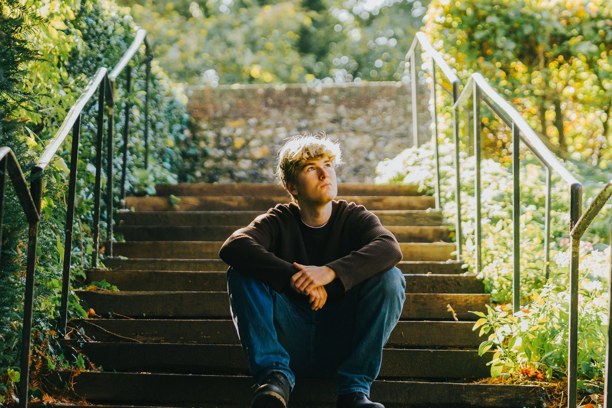 A young man with blond hair sitting on outdoor stairs surrounded by greenery, looking upward thoughtfully.