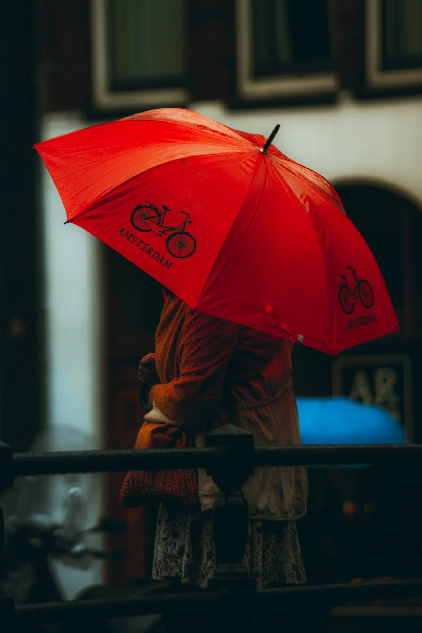Person holding a red umbrella with the word 'Amsterdam' and a bicycle graphic, standing in a dimly lit street.