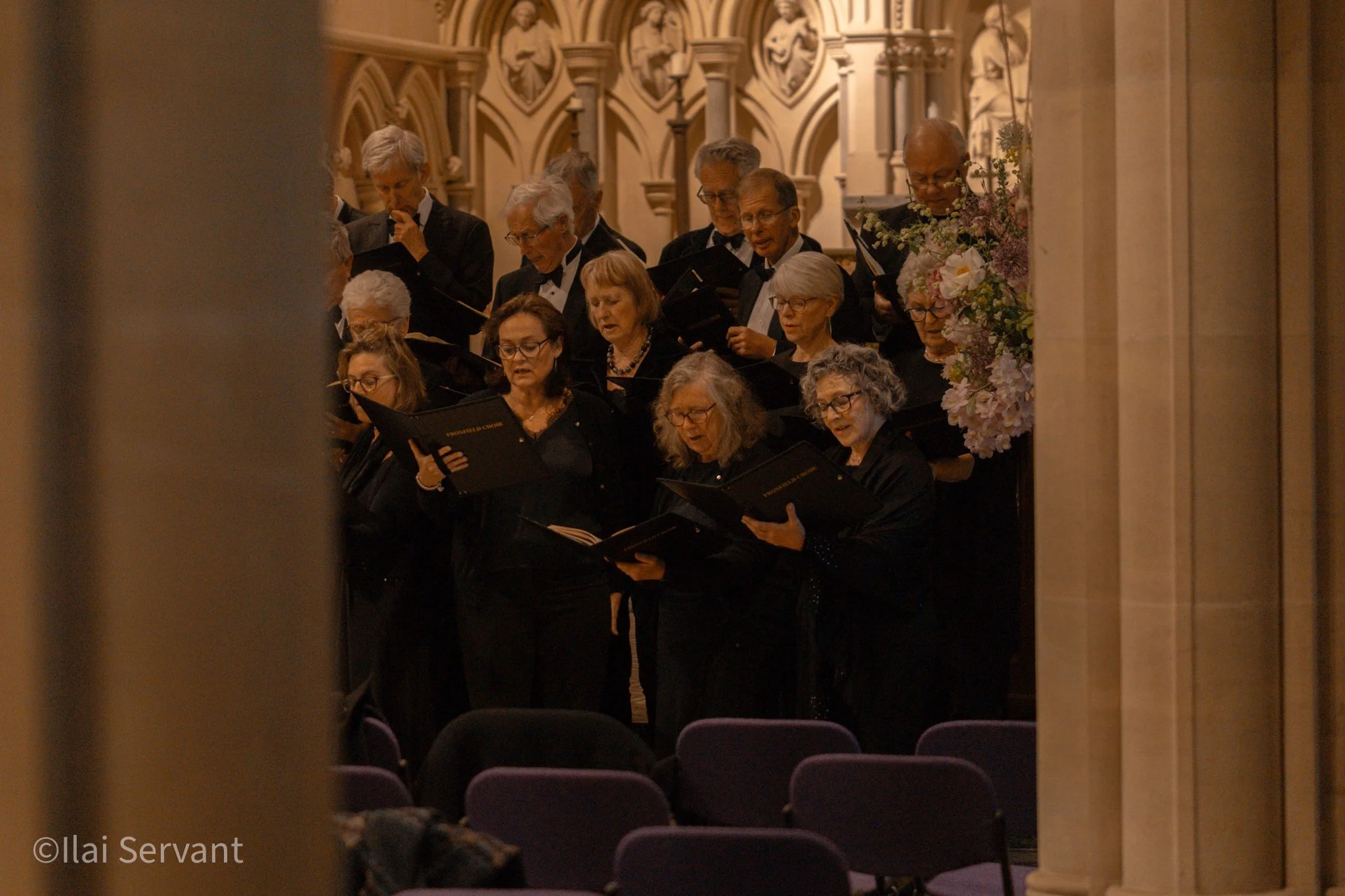 A choir of older men and women dressed in black, singing from black songbooks inside a church with intricate gothic architecture and stained glass windows, with purple chairs in the foreground.