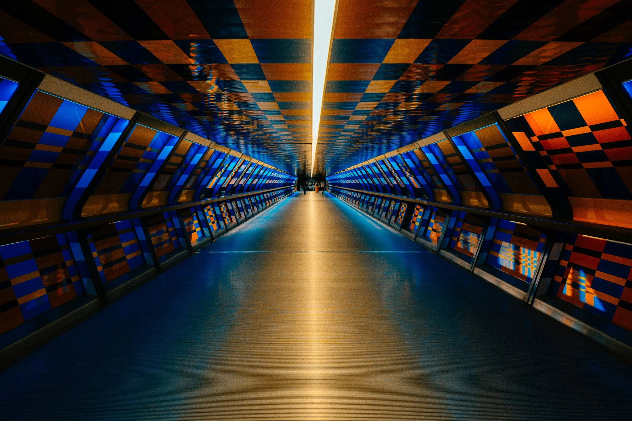 Indoor corridor with modern design, illuminated by a central strip light, featuring colorful geometric patterns on the walls and ceiling, with a few people visible at the end of the corridor.