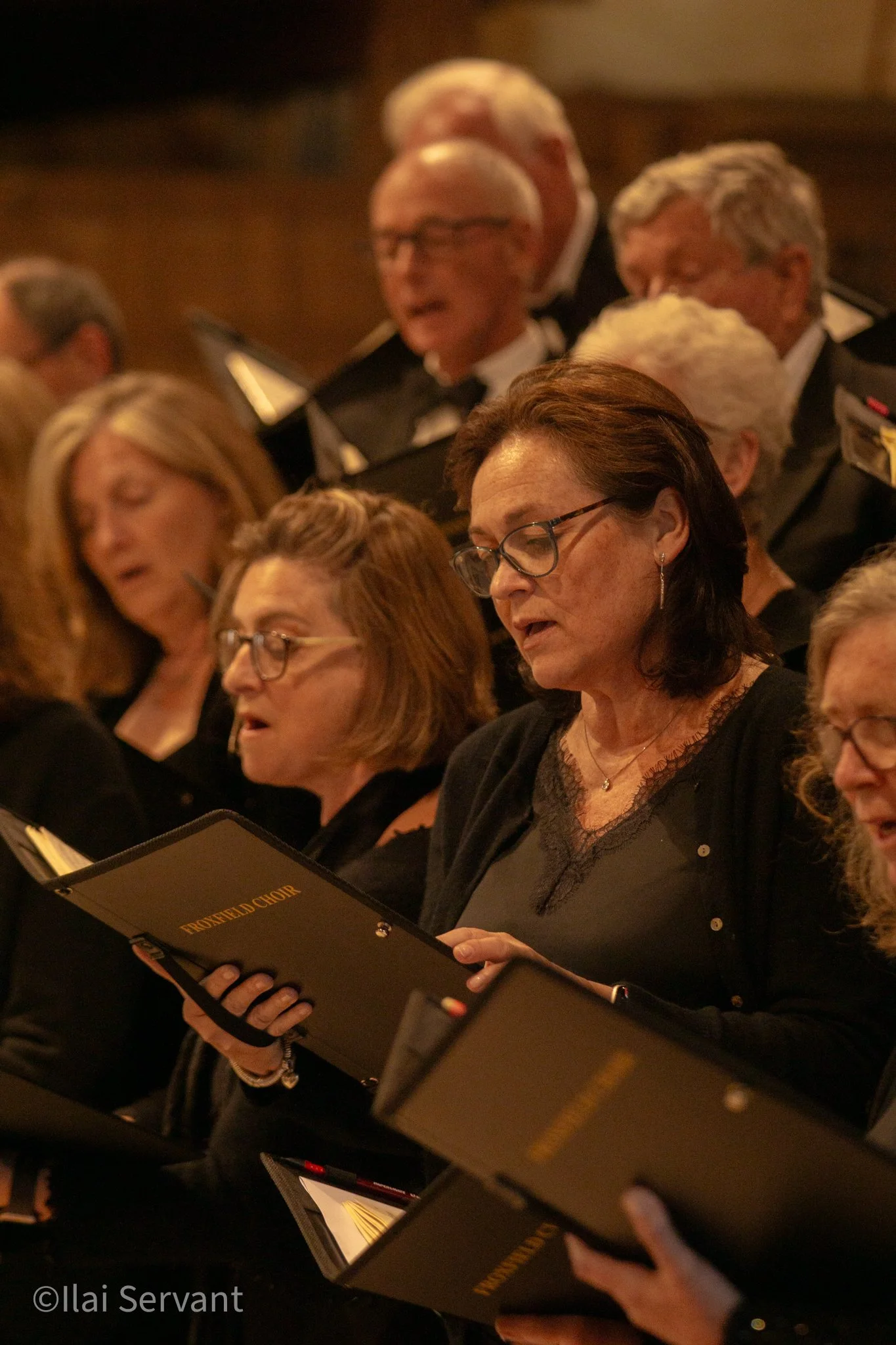 Group of adult women and men in choir robes singing and holding music books. The women are in the front and the men are in the back, with some wearing glasses.
