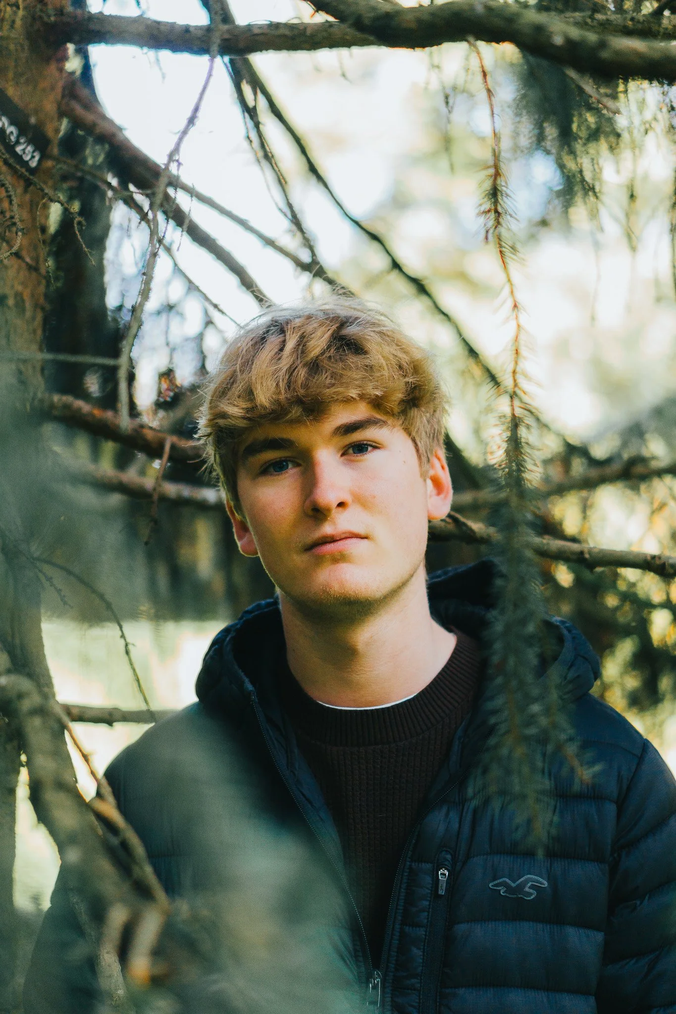 A young man with blond hair and blue eyes, wearing a dark jacket, stands outdoors among trees with blurred nature in the background.