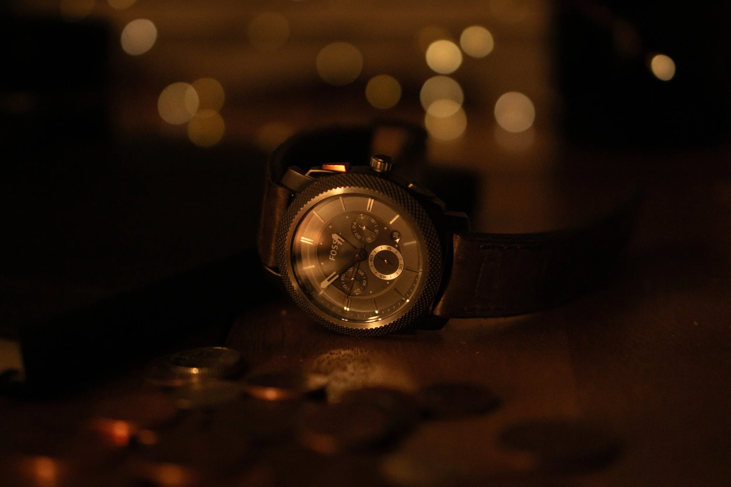 A Fossil brand wristwatch with a black leather strap, resting on a dark surface with scattered coins nearby, in a dimly lit setting with blurred bokeh lights in the background.