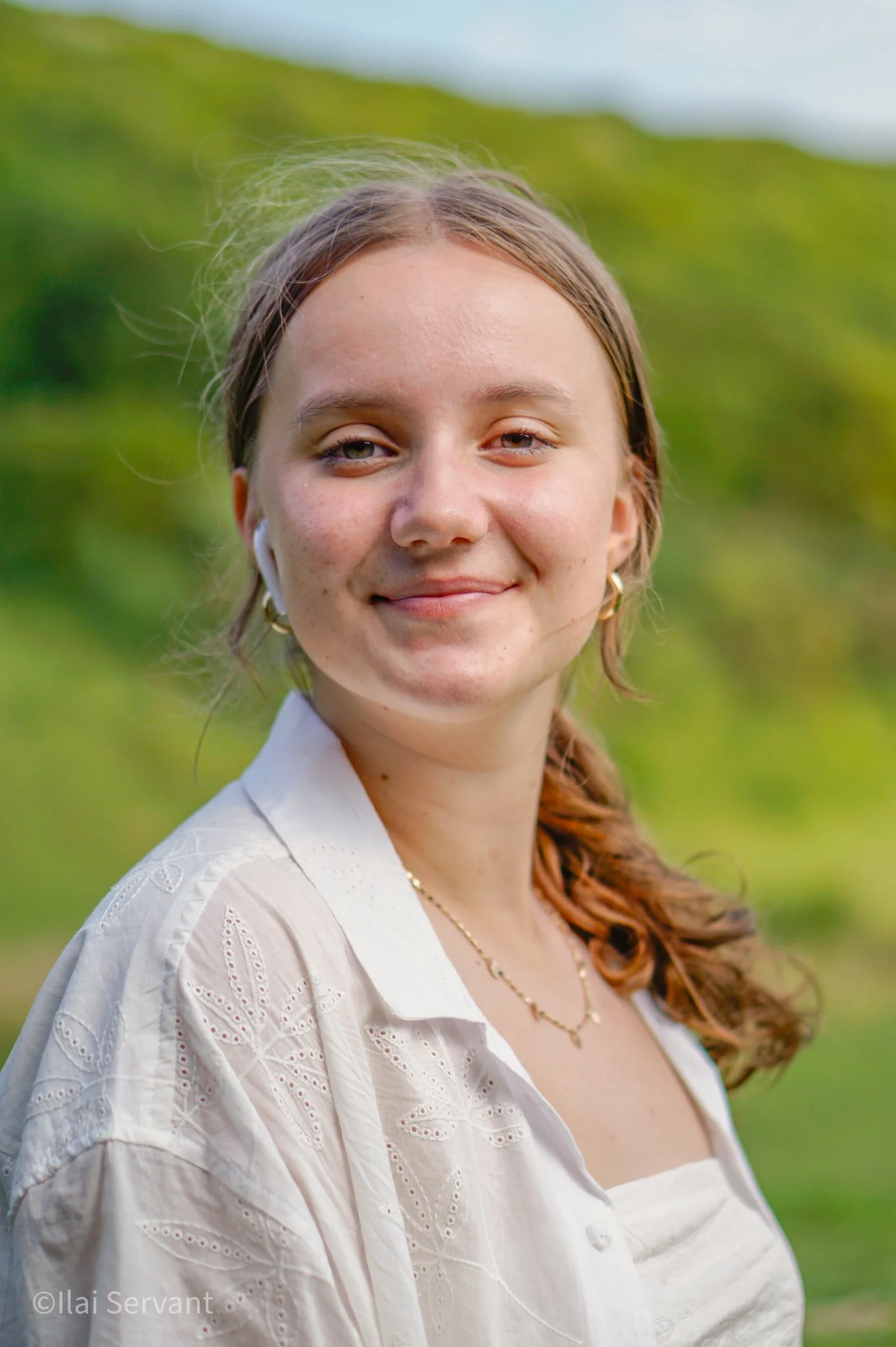 A young woman with light brown hair in a side braid, wearing earrings, a necklace, and a white embroidered blouse, smiling outdoors against a blurred green landscape background.