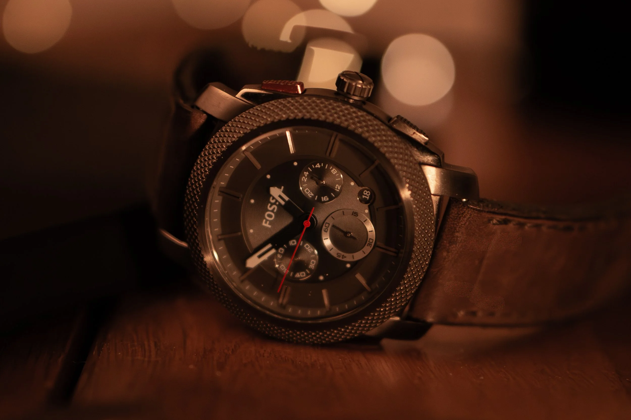A Fossil brand men's wristwatch with a black dial, multiple subdials, and a leather strap lying on a wooden surface, illuminated by warm ambient lighting with blurred lights in the background.