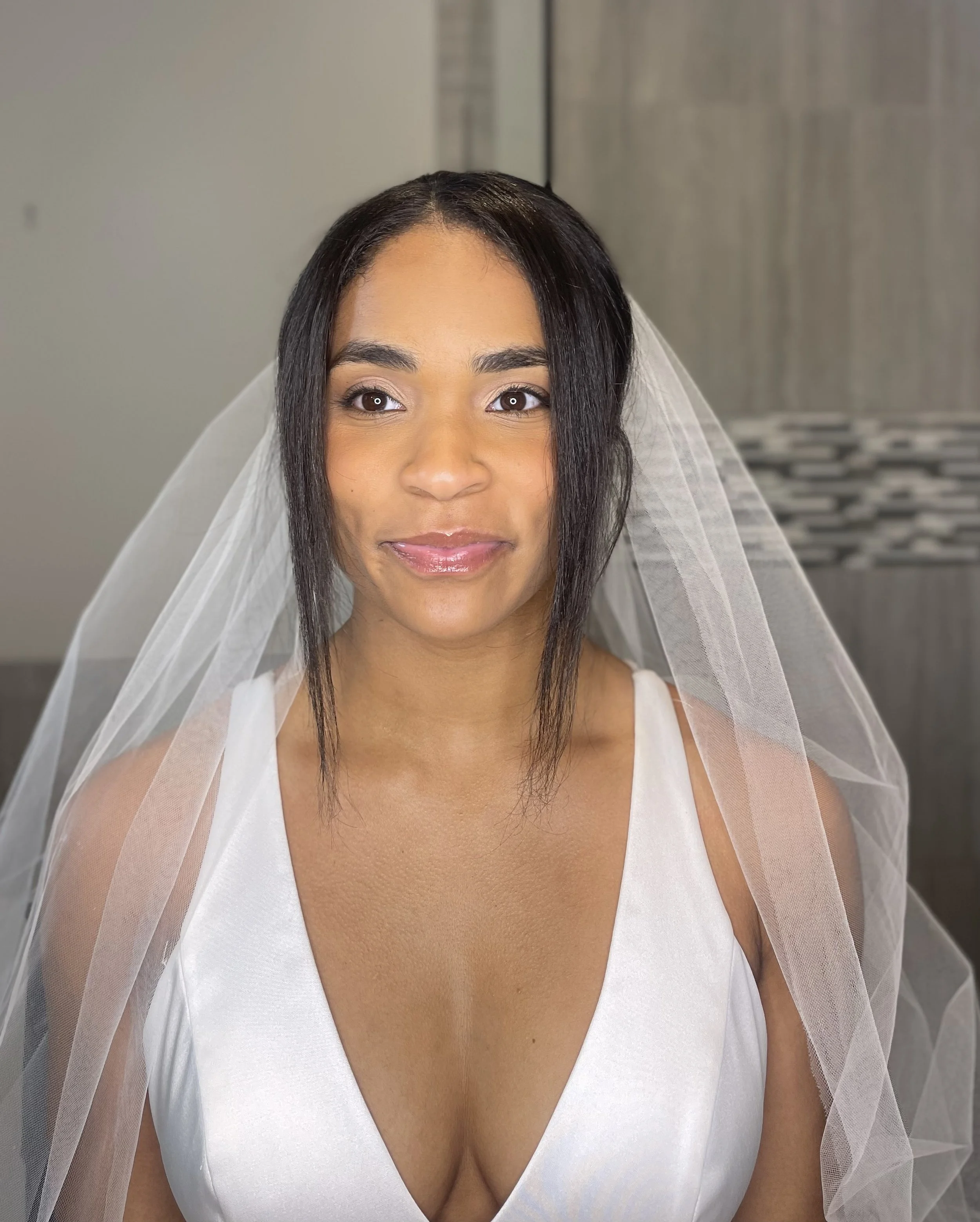 A woman dressed in a white sleeveless dress with a veil on her head, sitting indoors in front of a gray wall with a decorative tile section.