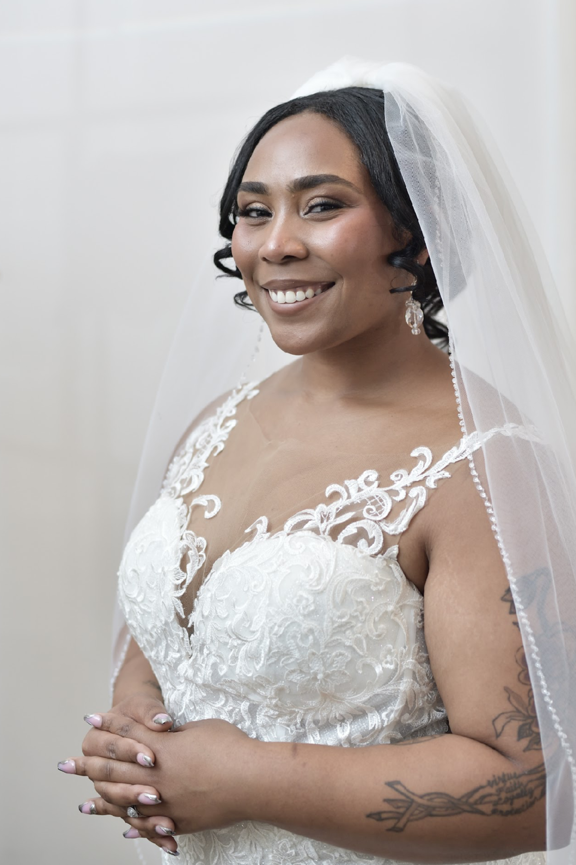 A woman in a wedding dress, smiling, wearing a veil and earrings, with tattoos on her arms.
