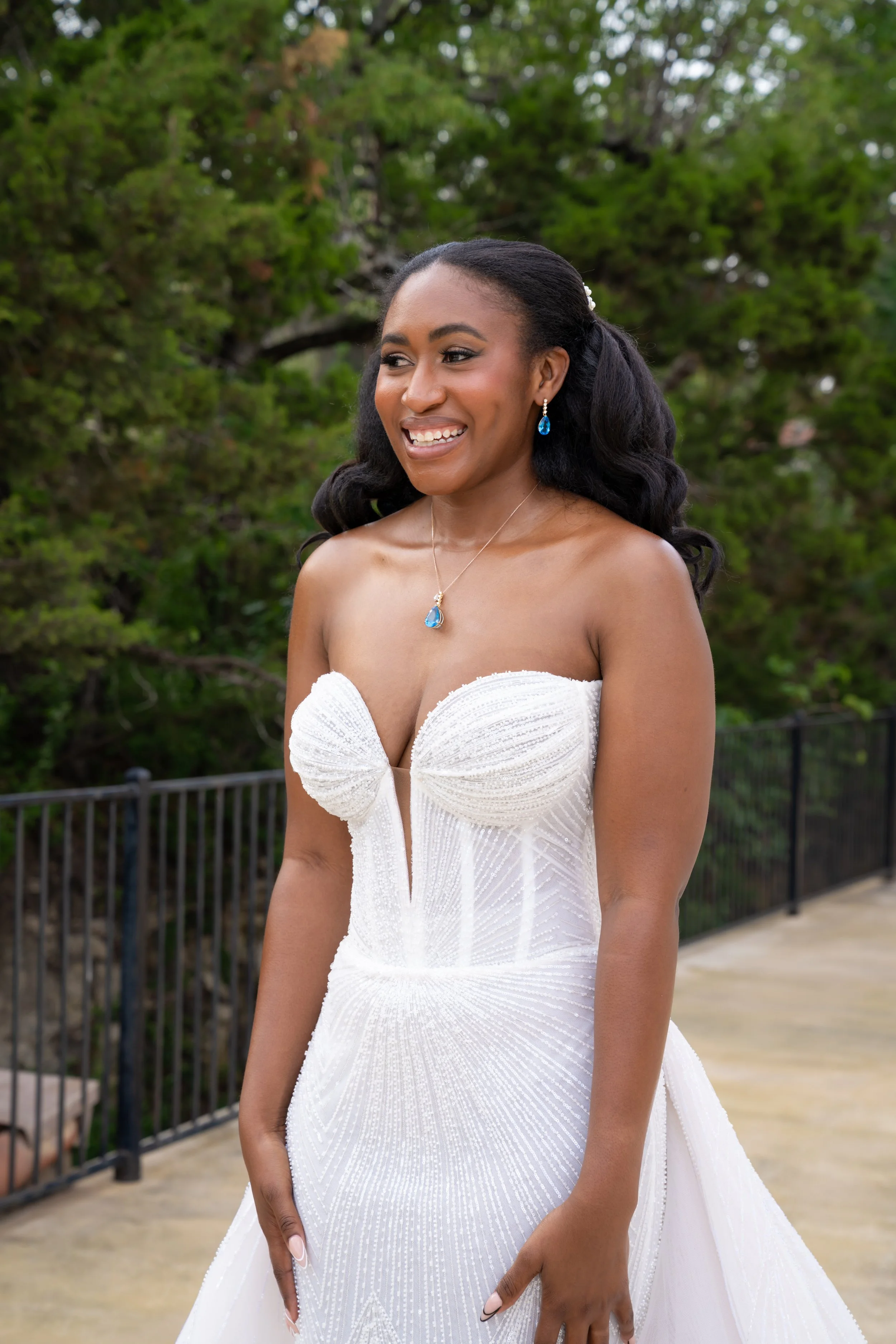 A smiling woman in a white strapless wedding dress with blue jewelry, standing outdoors with green trees in the background.