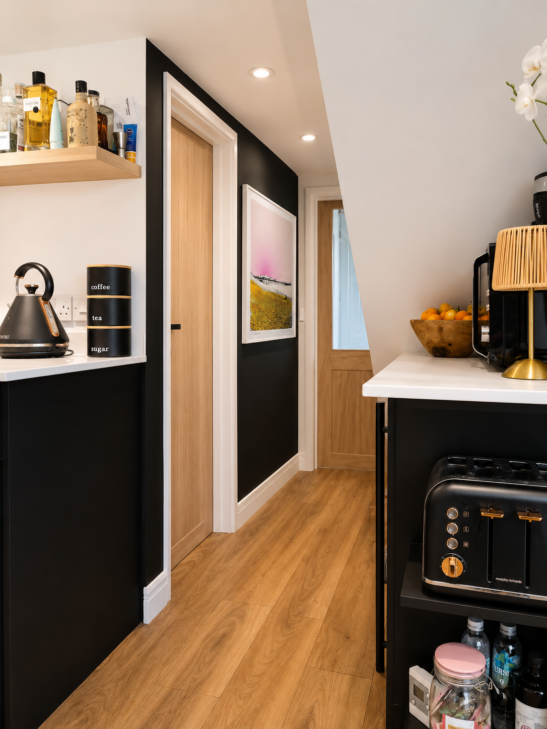 Dark kitchen hallway with matt black cabinets, white quartz worktop, bold pink landscape artwork and warm oak flooring, interior design Suffolk