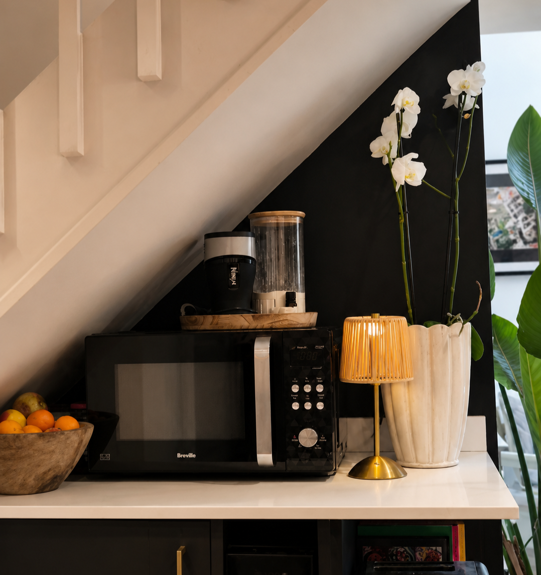 Kitchen counter vignette with matt black cabinets, brass lamp, white orchid and Breville microwave styled under staircase, Suffolk interior design