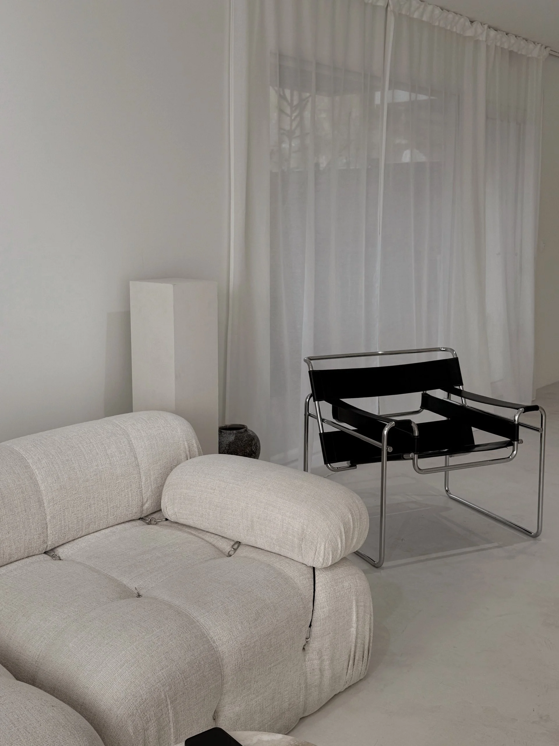 A living room with a light beige sofa, a black and chrome armchair, sheer white curtains, a tall white floor lamp, and a small dark vase on the floor.