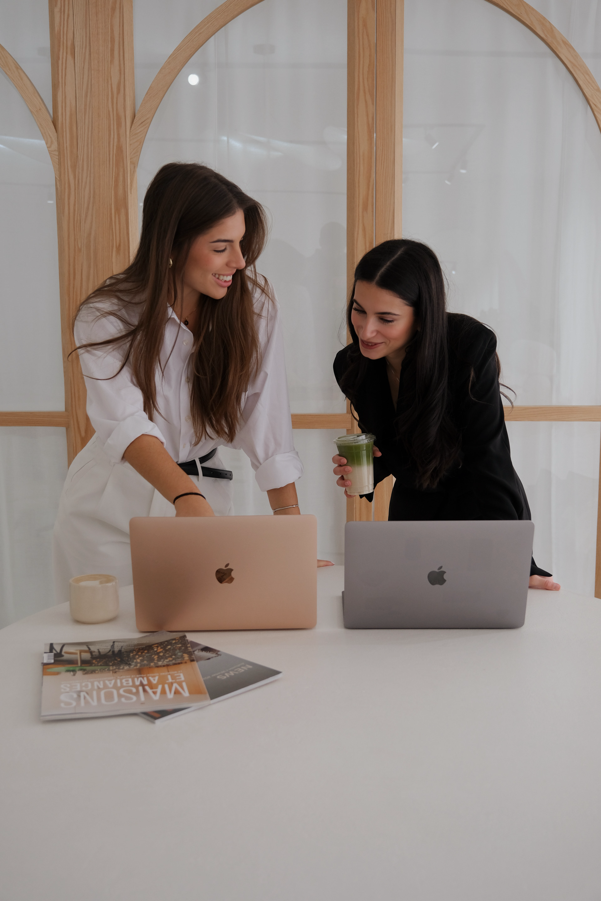 Two women standing at a white table with laptops, magazines, a candle, and a drink, smiling and engaging in a conversation in a modern office setting.