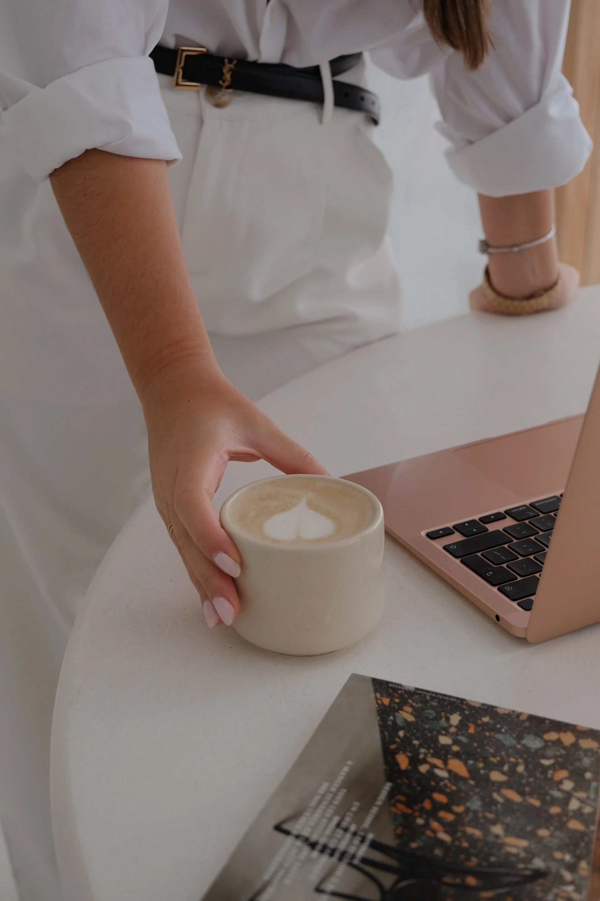 Person in white shirt and light-colored pants holding a cup of coffee with latte art, standing next to a pink laptop on a white table.