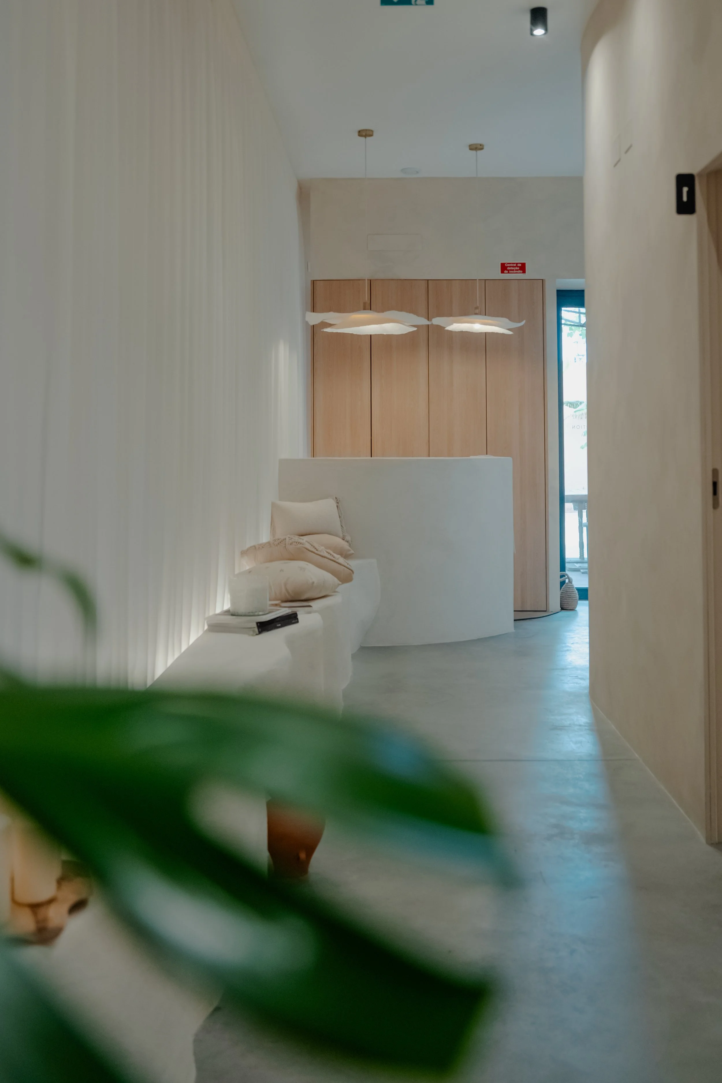 Minimalist interior with curved white sofa, beige pillows, wooden cabinets, and modern suspended white light fixtures in a bright room.