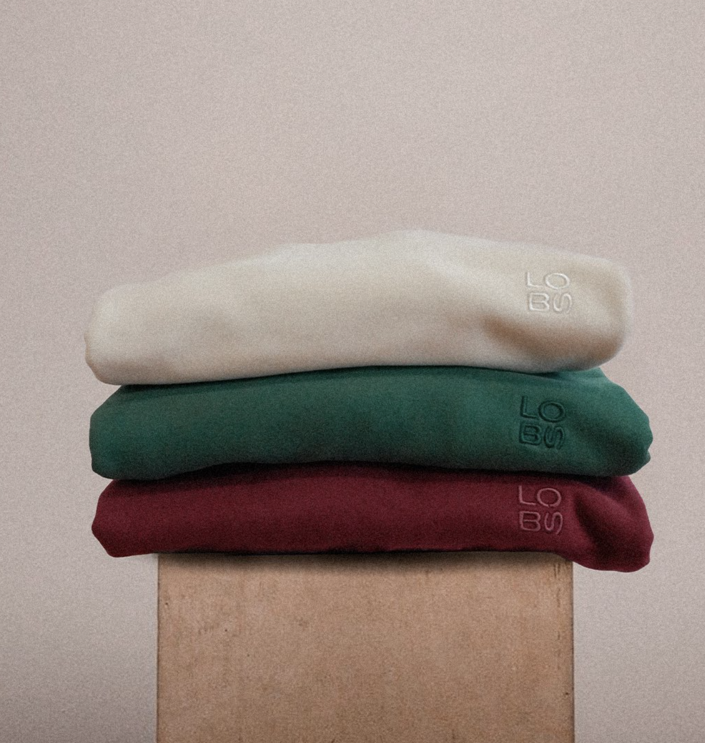Stack of three folded fabric clothes in white, green, and red on a beige block against a plain background.
