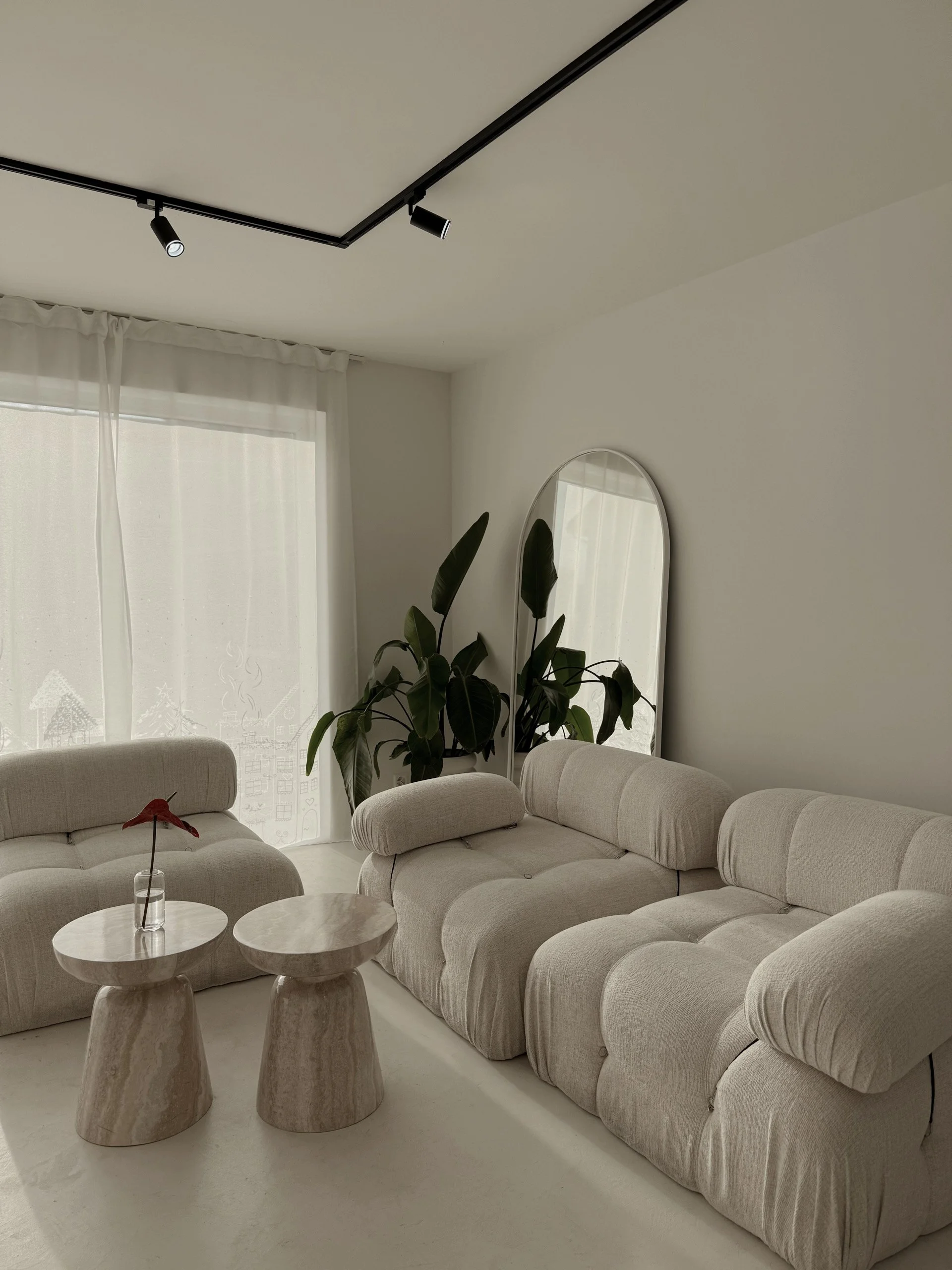Minimalist living room with beige sectional sofa, two matching marble side tables, a tall potted plant, a mirror, and sheer curtains on a window.