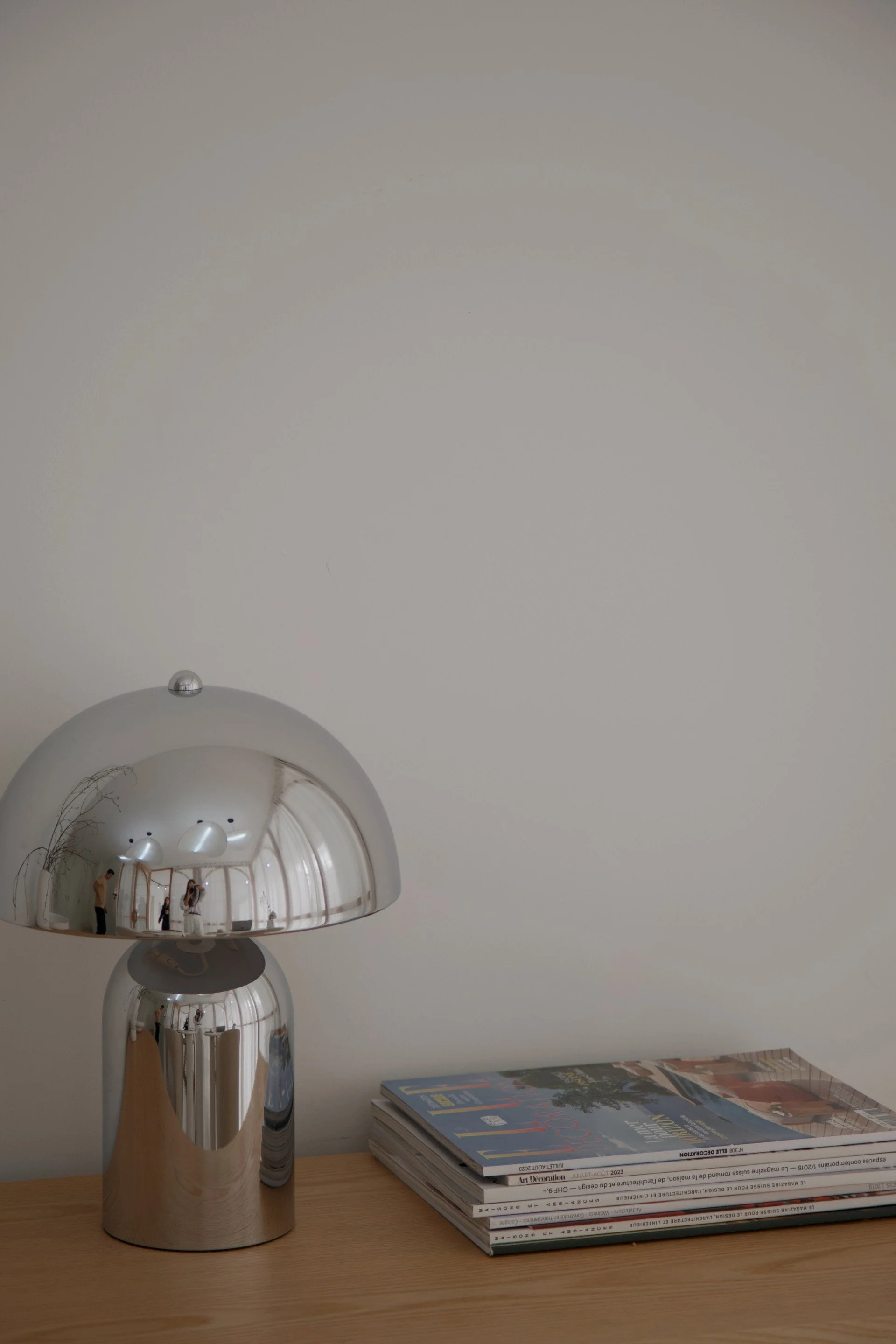 A modern silver mushroom-shaped lamp on a wooden surface, accompanied by a stack of magazines.