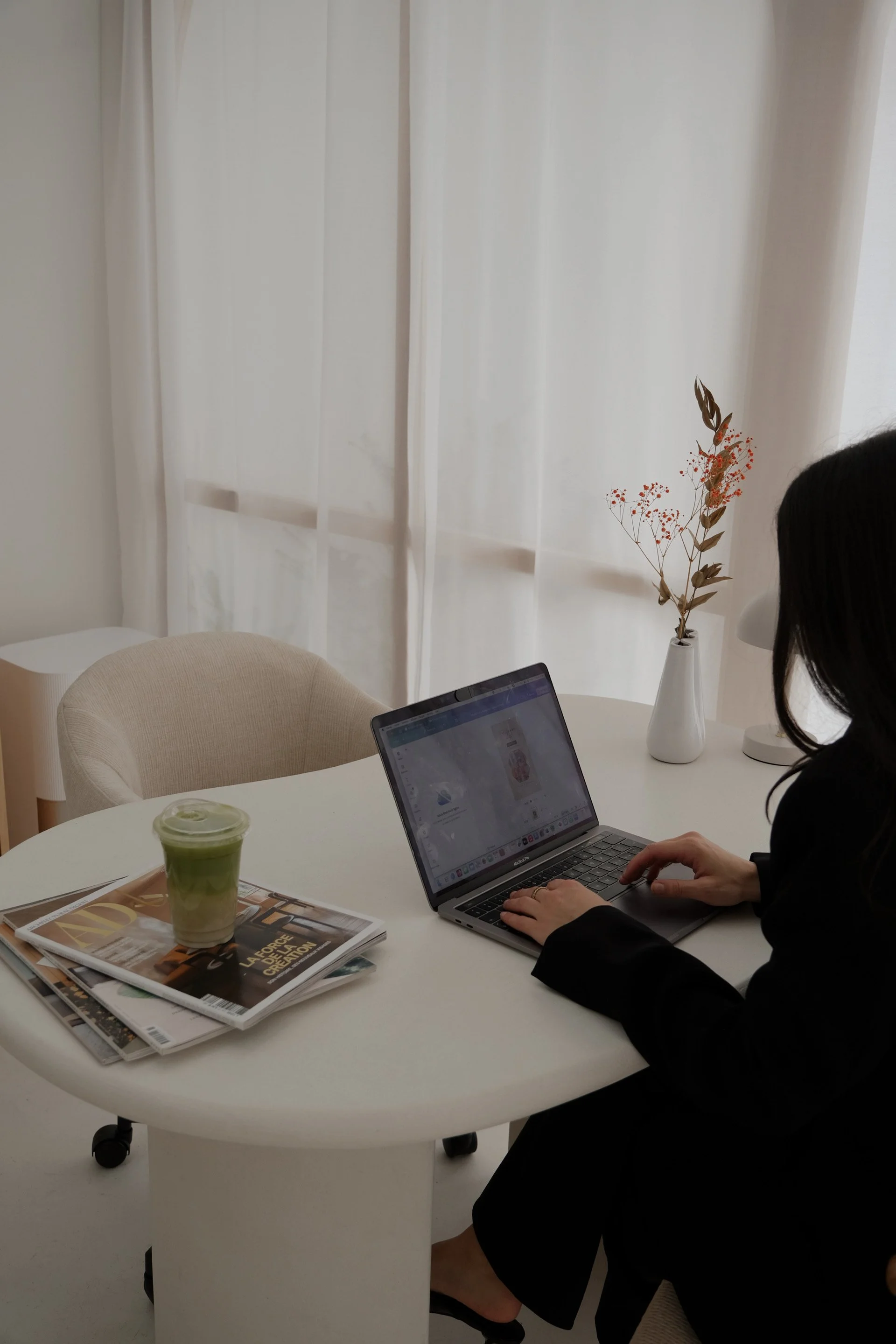 A woman sitting at a white table using a laptop, with a green drink and magazines on the table, in a room with white curtains and a vase with dried flowers.