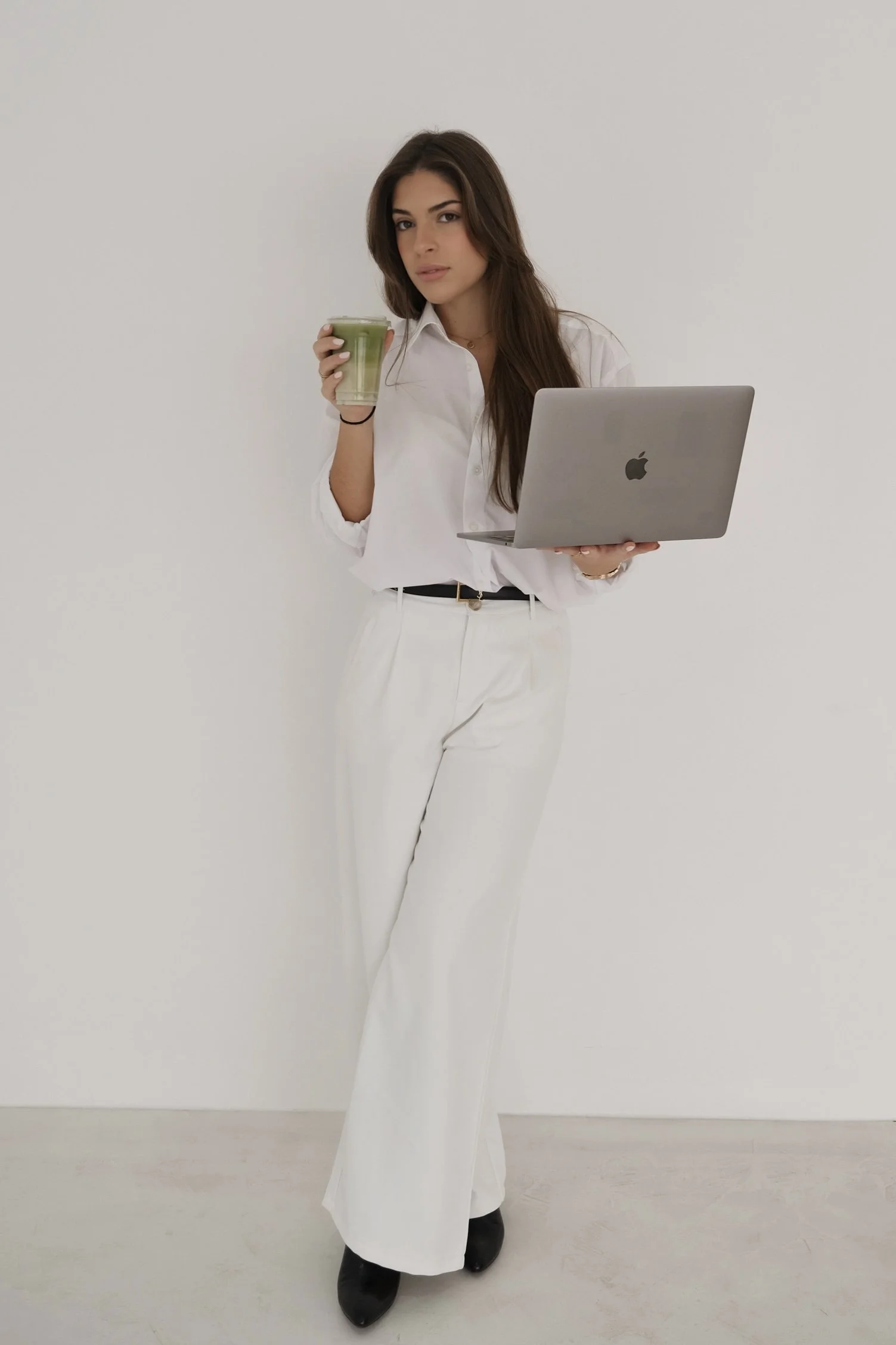 A young woman with long brown hair, wearing a white blouse and white wide-leg pants, holding a laptop in one hand and a green smoothie in the other, standing against a plain white wall.
