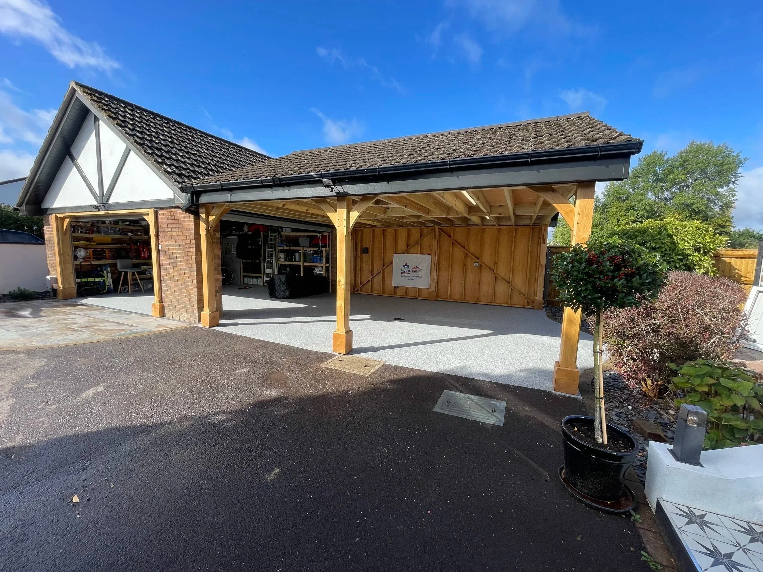Residential garage with wooden extension, partially open doors, and a paved driveway. Storage shelves and equipment visible inside, some plants and shrubs outside.