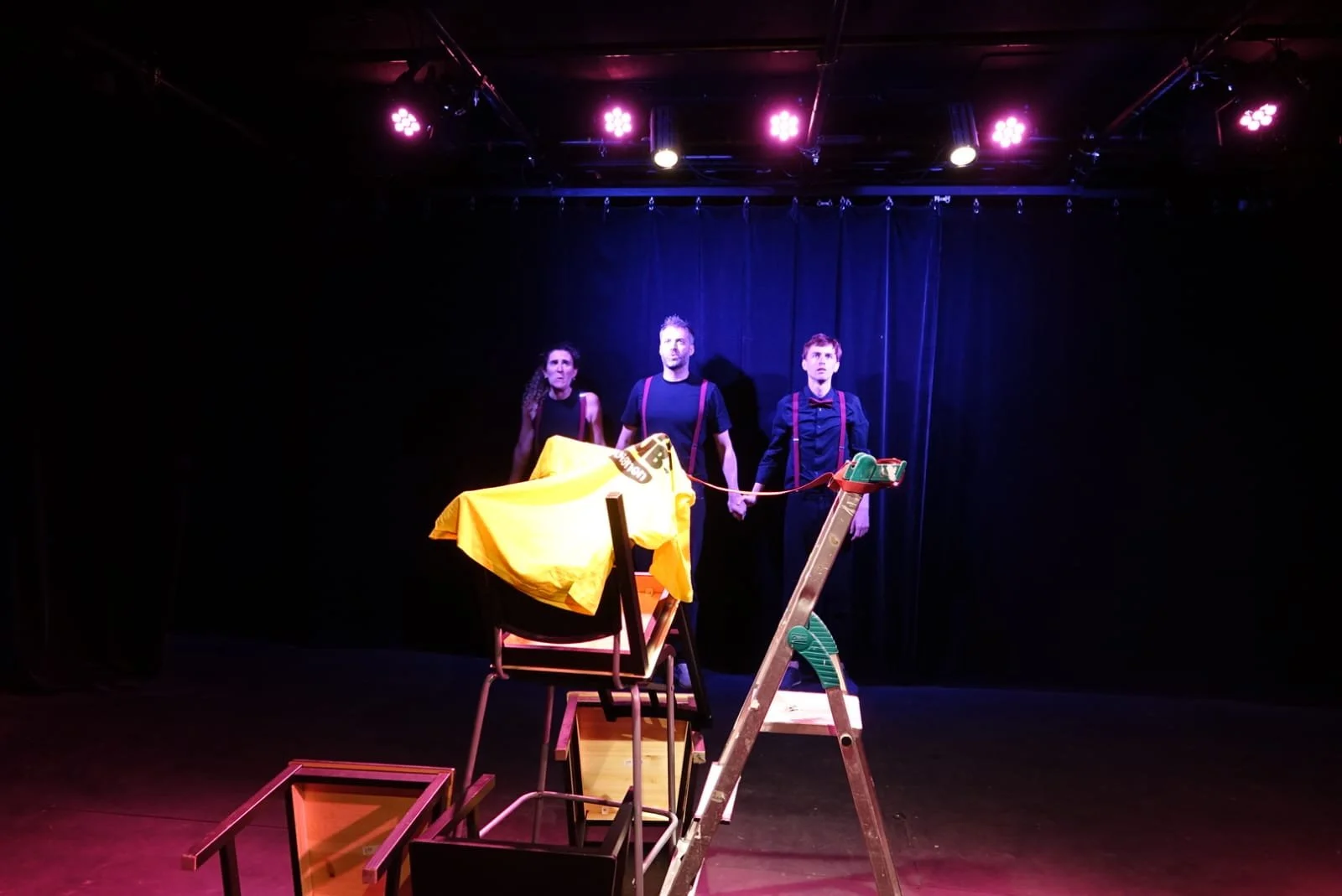 Scène de théâtre avec trois personnes en costume, un éclairage violet, des chaises, un escabeau, et une scène sombre en arrière-plan.