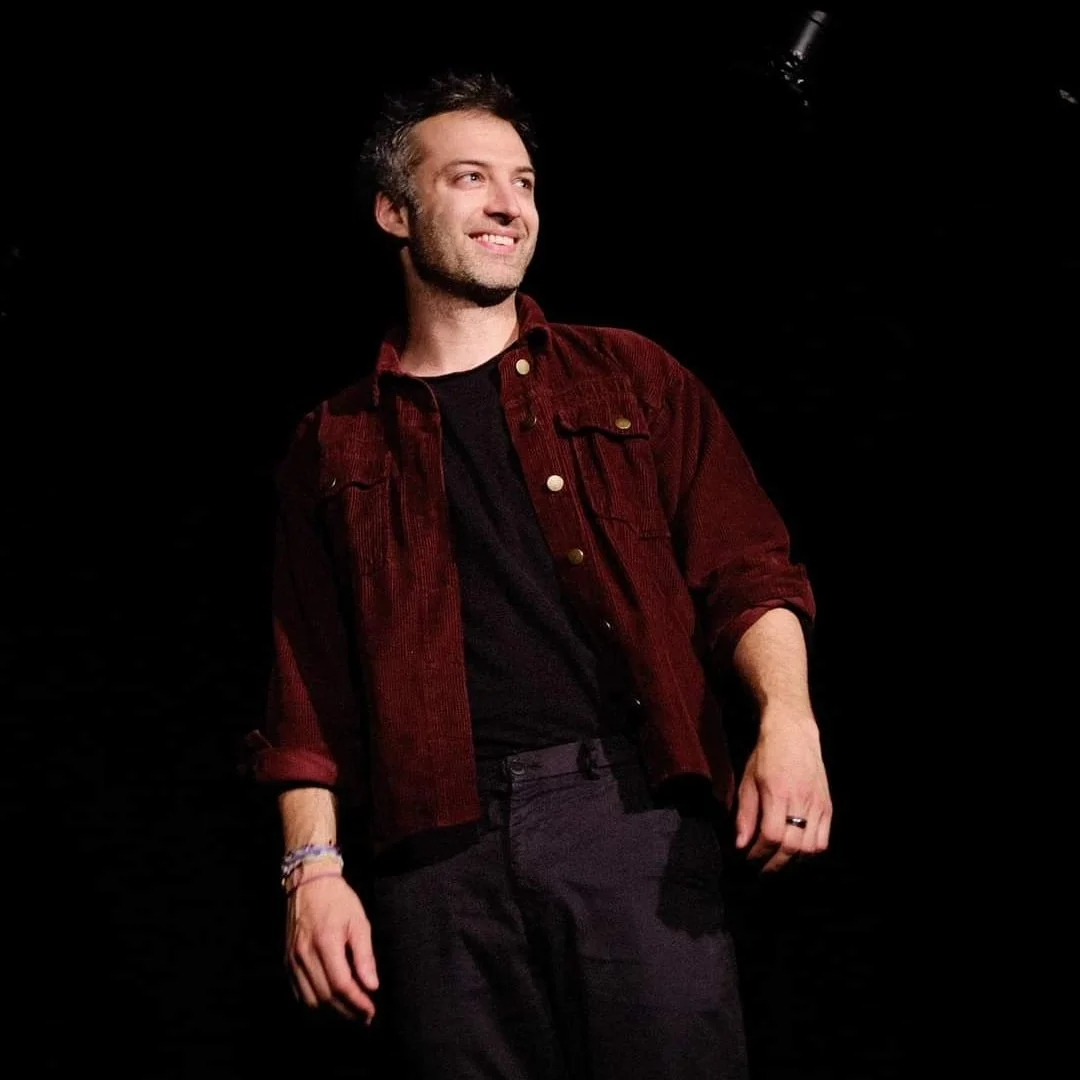 Paul improvise. Un homme souriant avec une barbe porte une veste en velours bordeaux et un t-shirt noir, il est sur scène avec un fond noir.