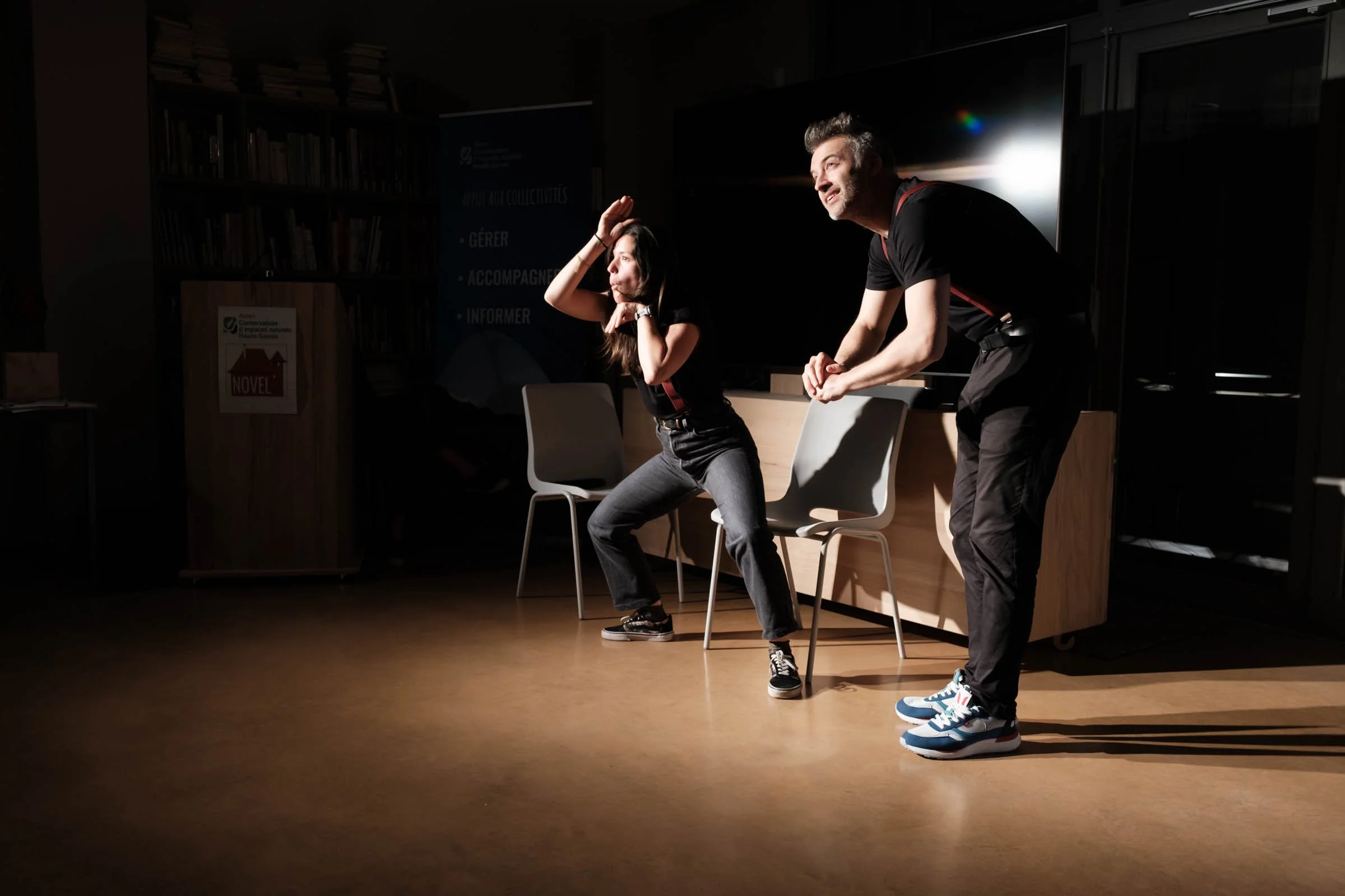 Deux personnes, un homme et une femme, en train de faire une représentation théâtrale ou un spectacle improvisé dans une pièce sombre, avec des chaises, un meuble en bois et des étagères remplies de livres en arrière-plan.