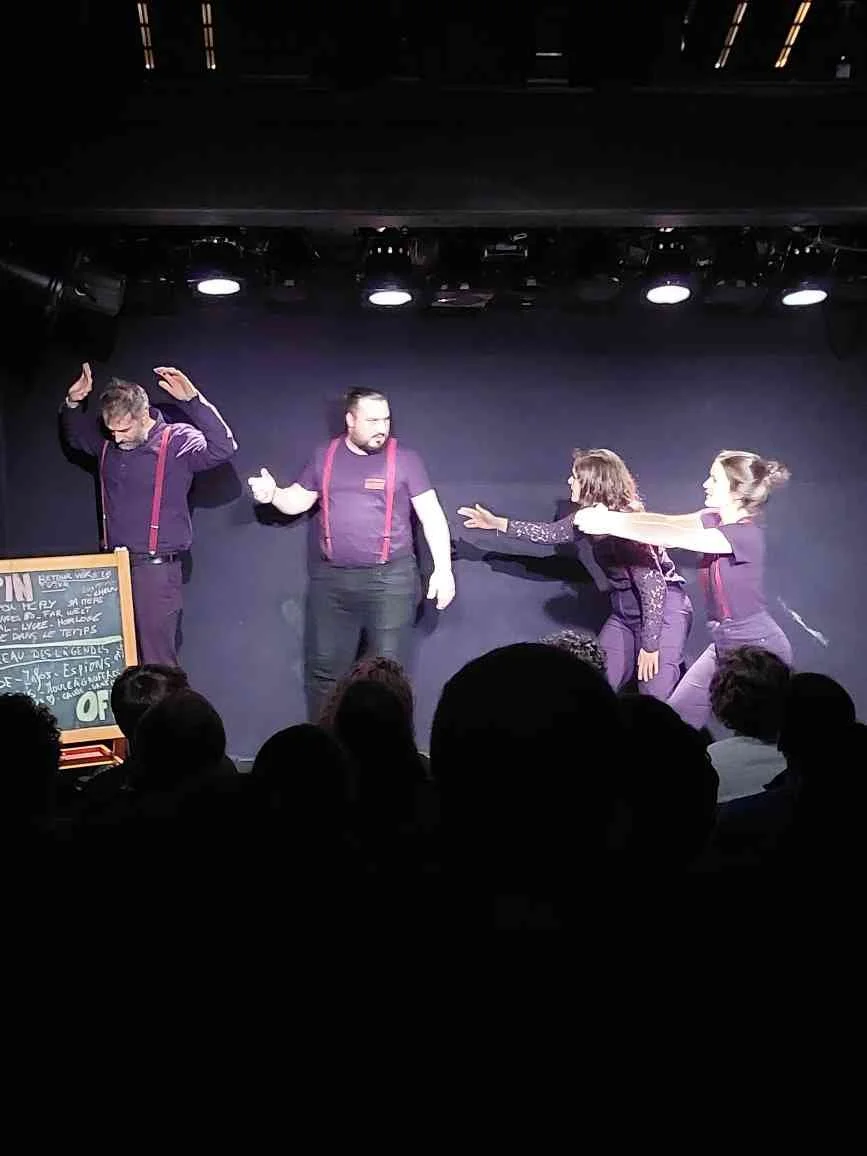 Quatre personnes sur scène, deux hommes et deux femmes, apparentés à une performance ou une pièce de théâtre, avec un tableau noir à gauche.